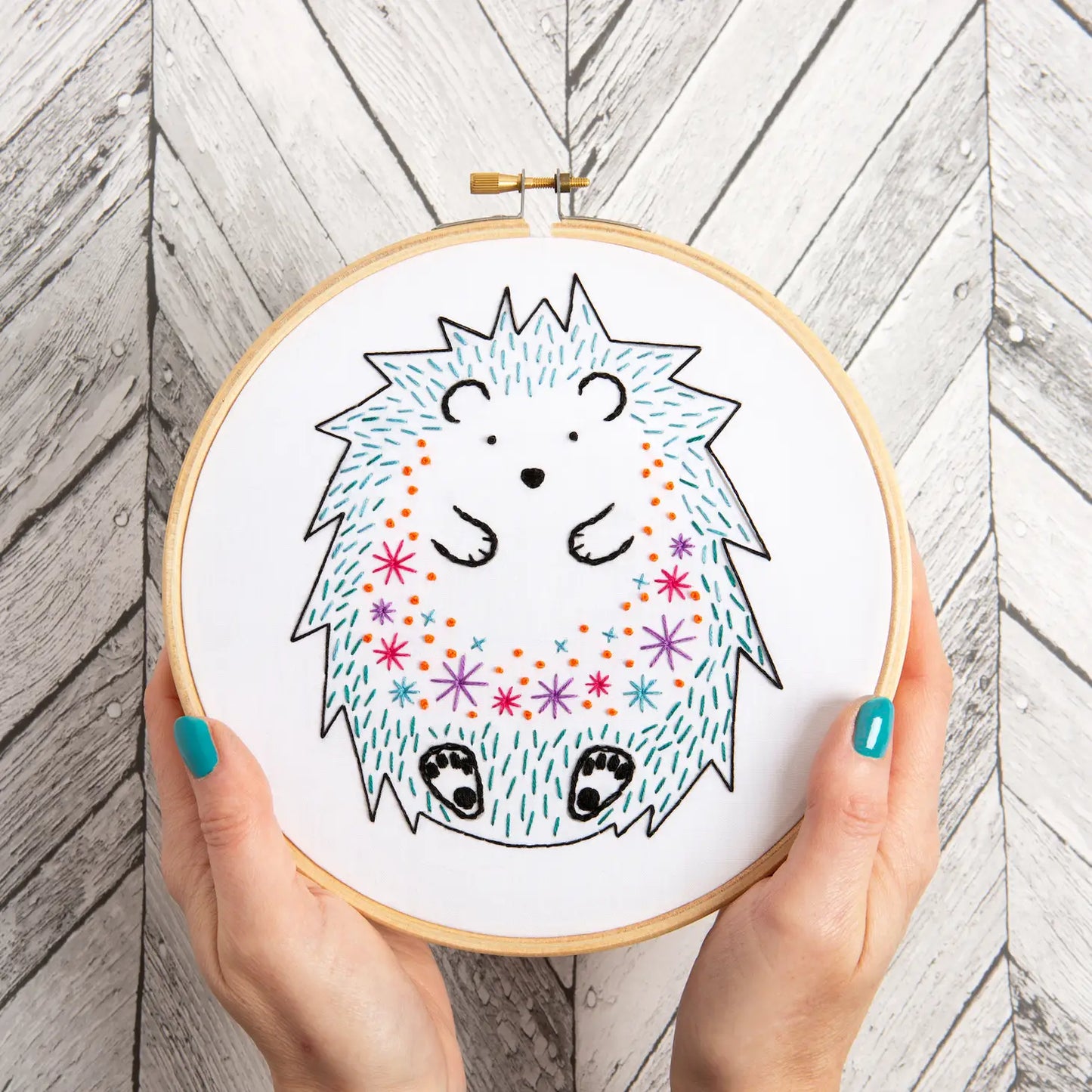 Embroidery hoop with a colorful hedgehog design held by hands on a wooden background