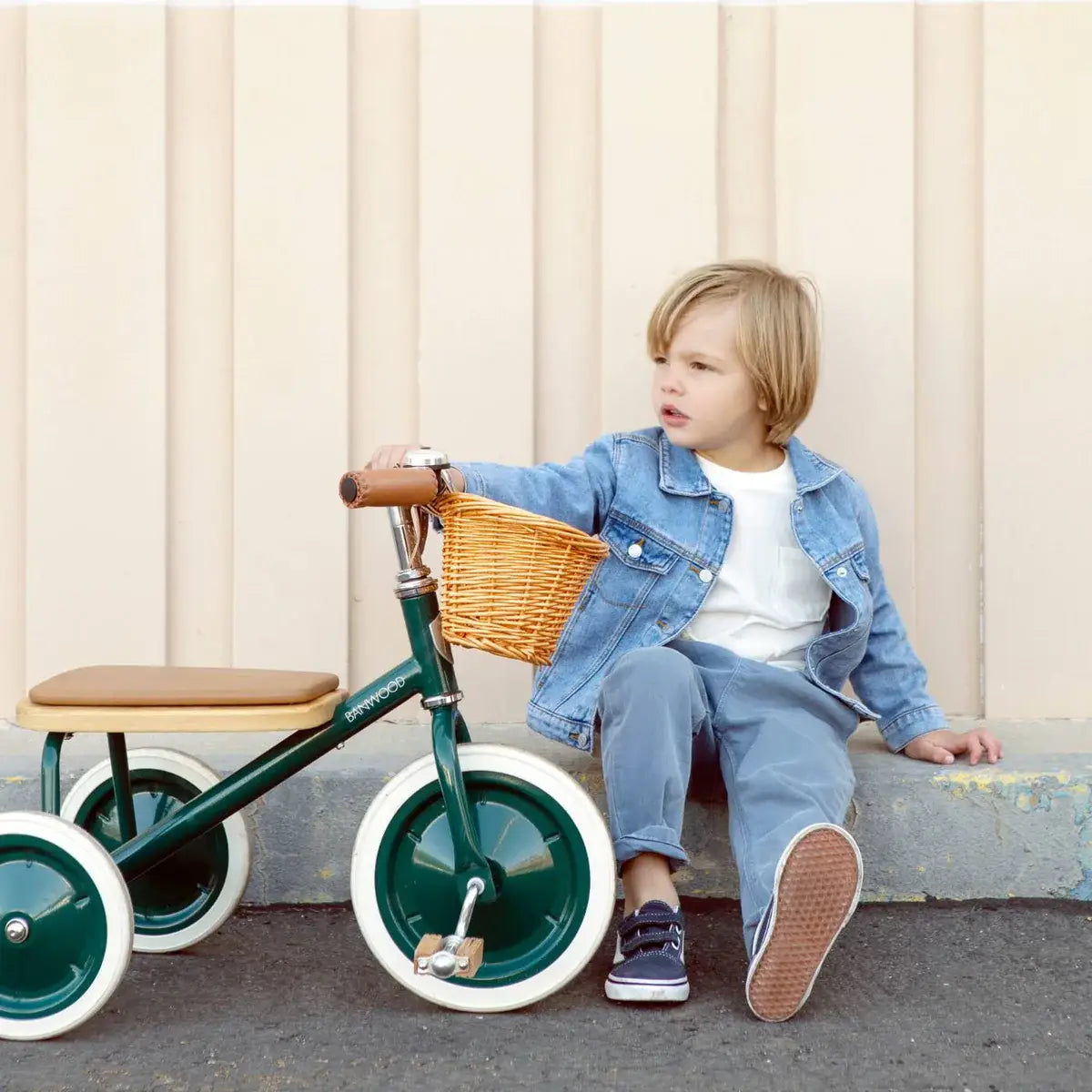 Child sitting on a tricycle with a wicker basket against a beige wall.