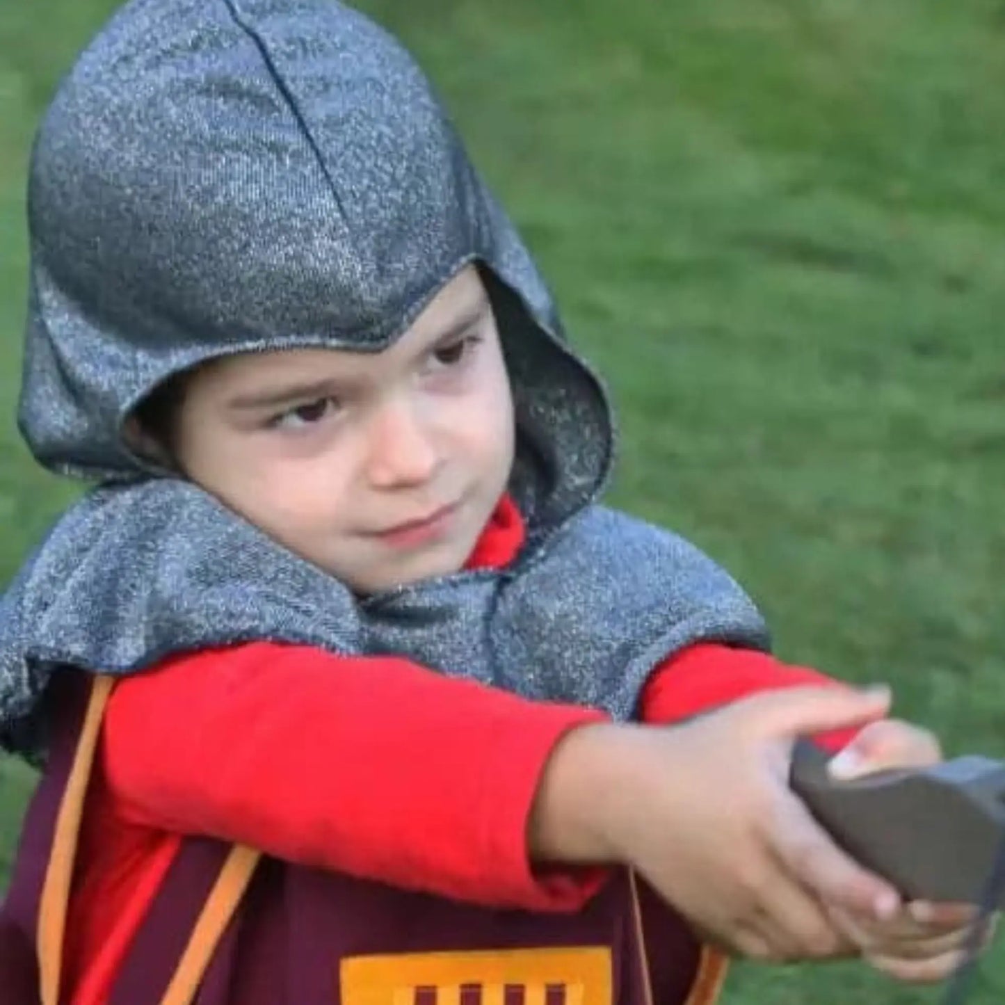 Child in a knight's costume with a chainmail hood and red sleeves, holding a toy sword outdoors.