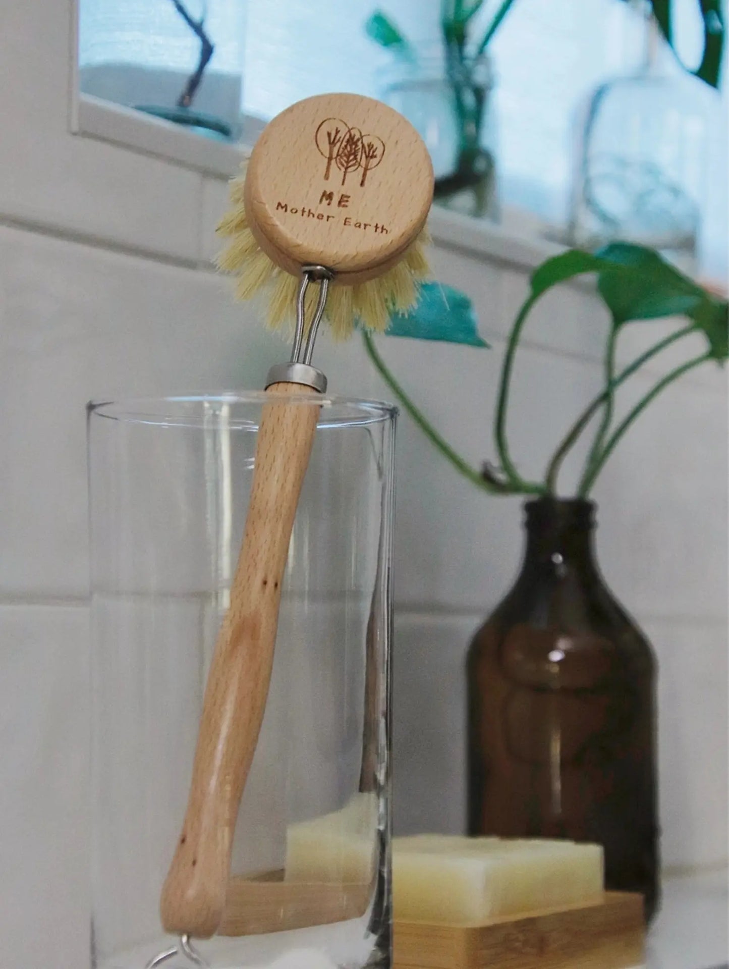 Cleaning brush with wooden handle and 'Mother Earth' branding, placed inside a glass container with a bar of soap on a tiled bathroom counter.
