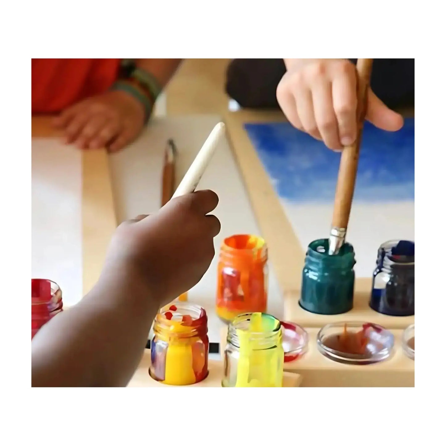 Children's hands with paintbrushes and colorful paint containers on a table.