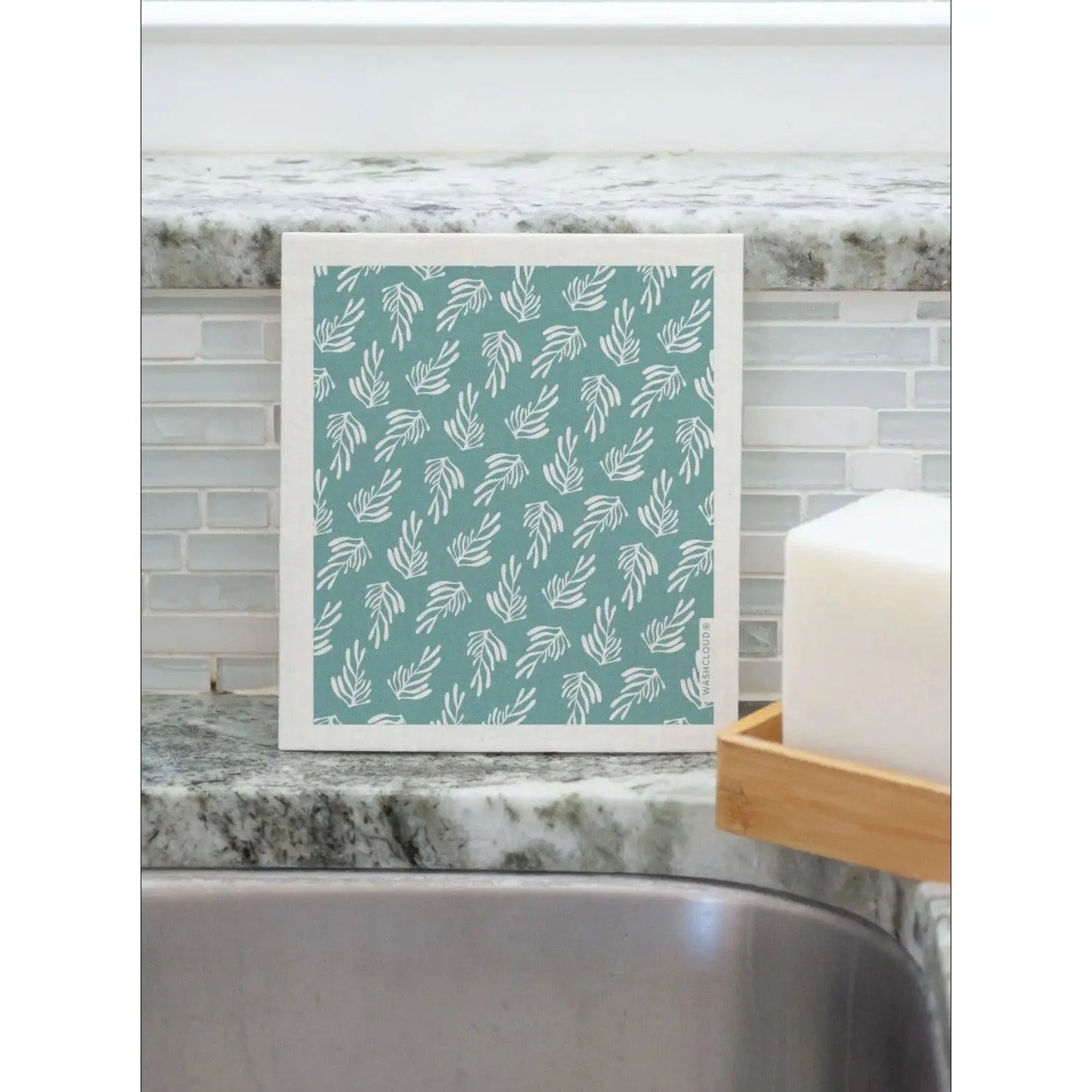 Swedish Dishcloth in Teal and White with Kelp Design Propped up against the kitchen sink backsplash