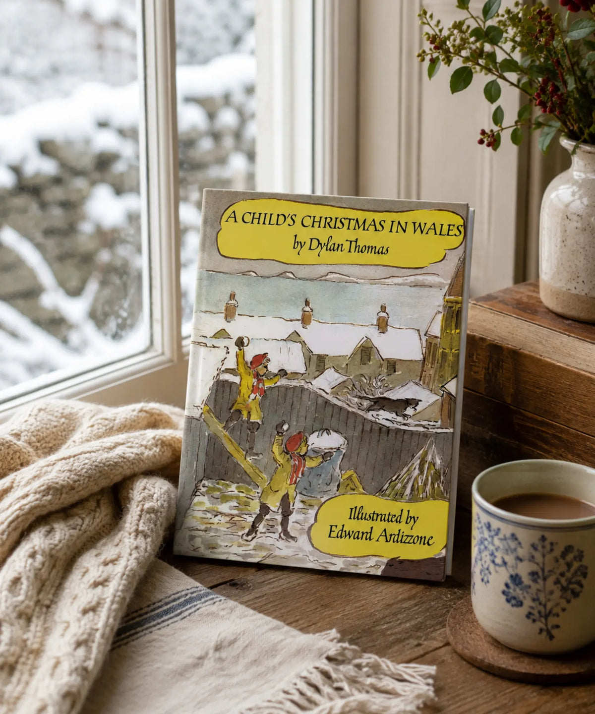 Book titled 'A Child's Christmas in Wales' by Dylan Thomas on a windowsill with a cup of coffee and blanket.