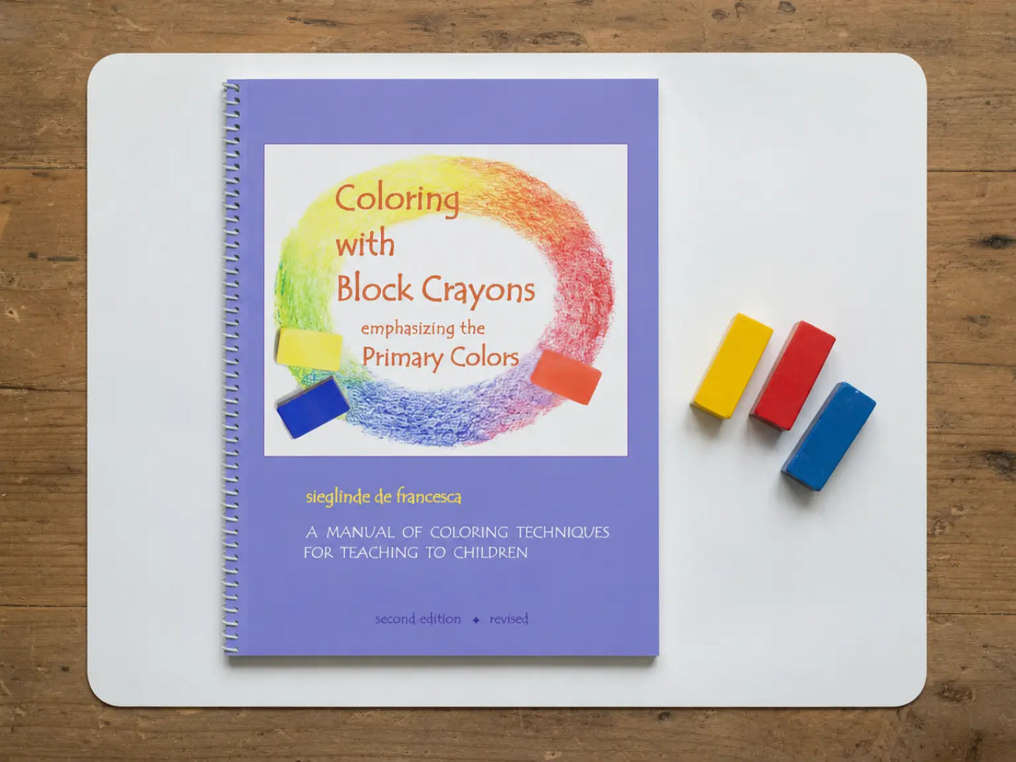 A spiral-bound book "Coloring with Block Crayons" on a white board over a wooden surface, with three primary color blocks in yellow, red, and blue.
