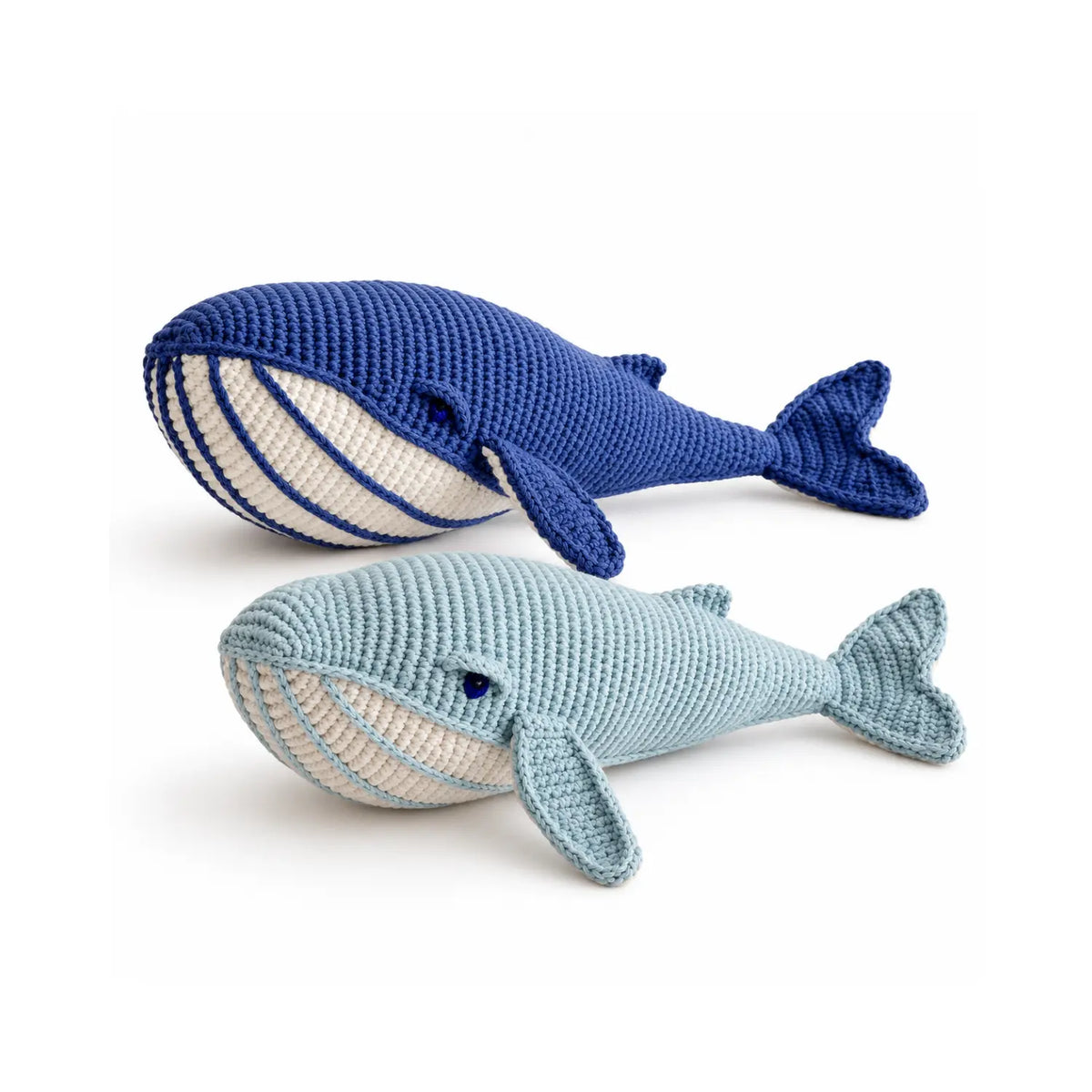 Two organic cotton crochet whale soft toys side by side, one in deep blue and one in misty blue, each with a cream striped belly