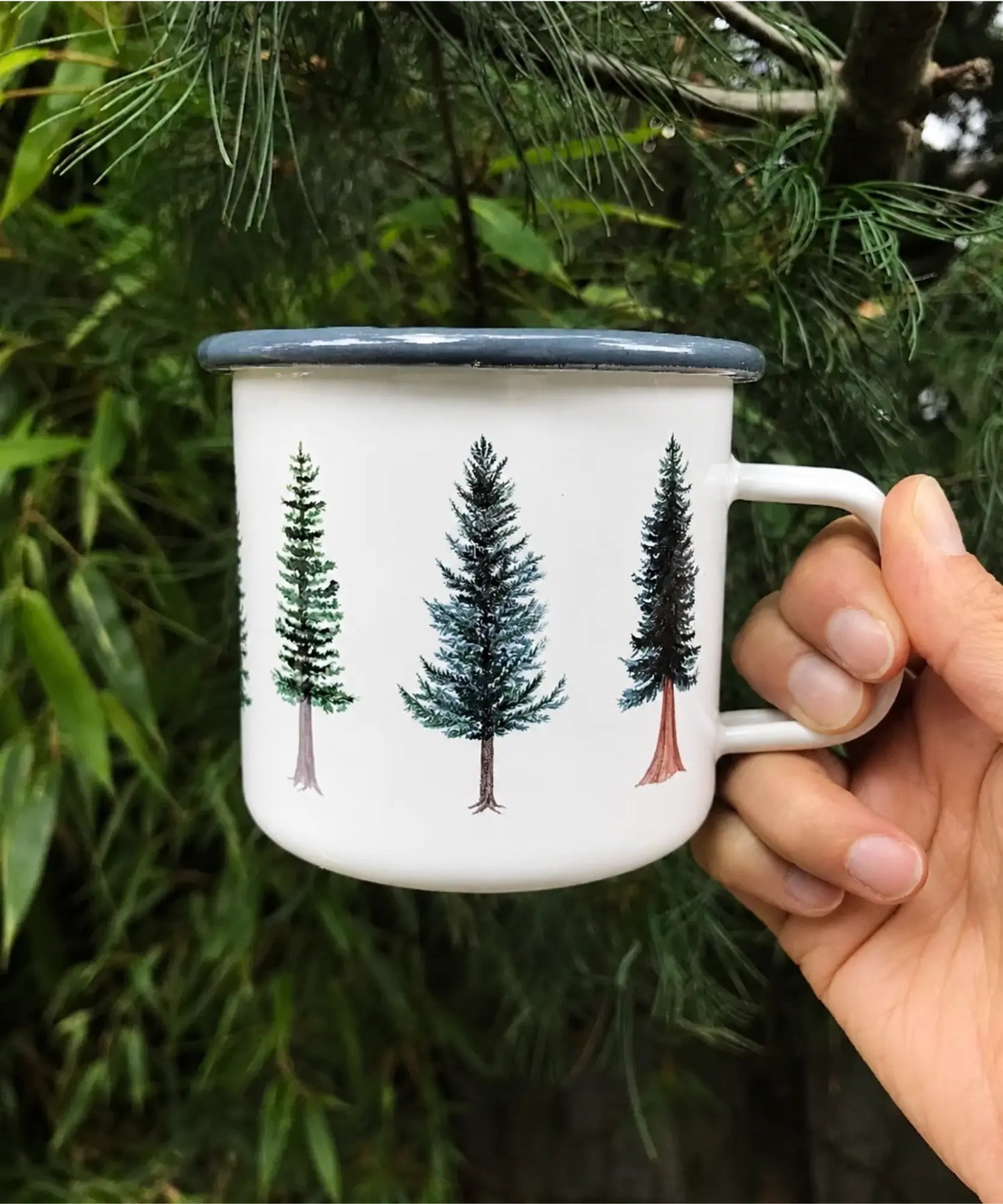 White enameled camping mug with evergreen tree design held in front of green foliage