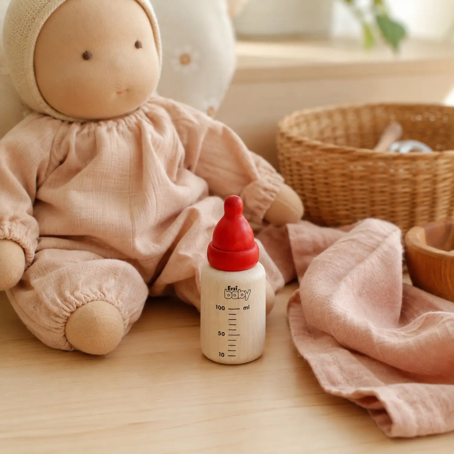 A wooden baby bottle with a red nipple and white measuring markings, placed next to a doll in pink