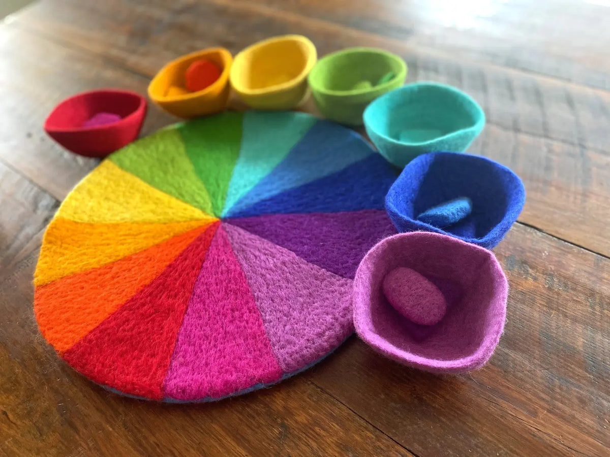 Rainbow felt bowls arranged in color order around a 12-color wool felt play mat on a wooden table, with felt pieces inside.