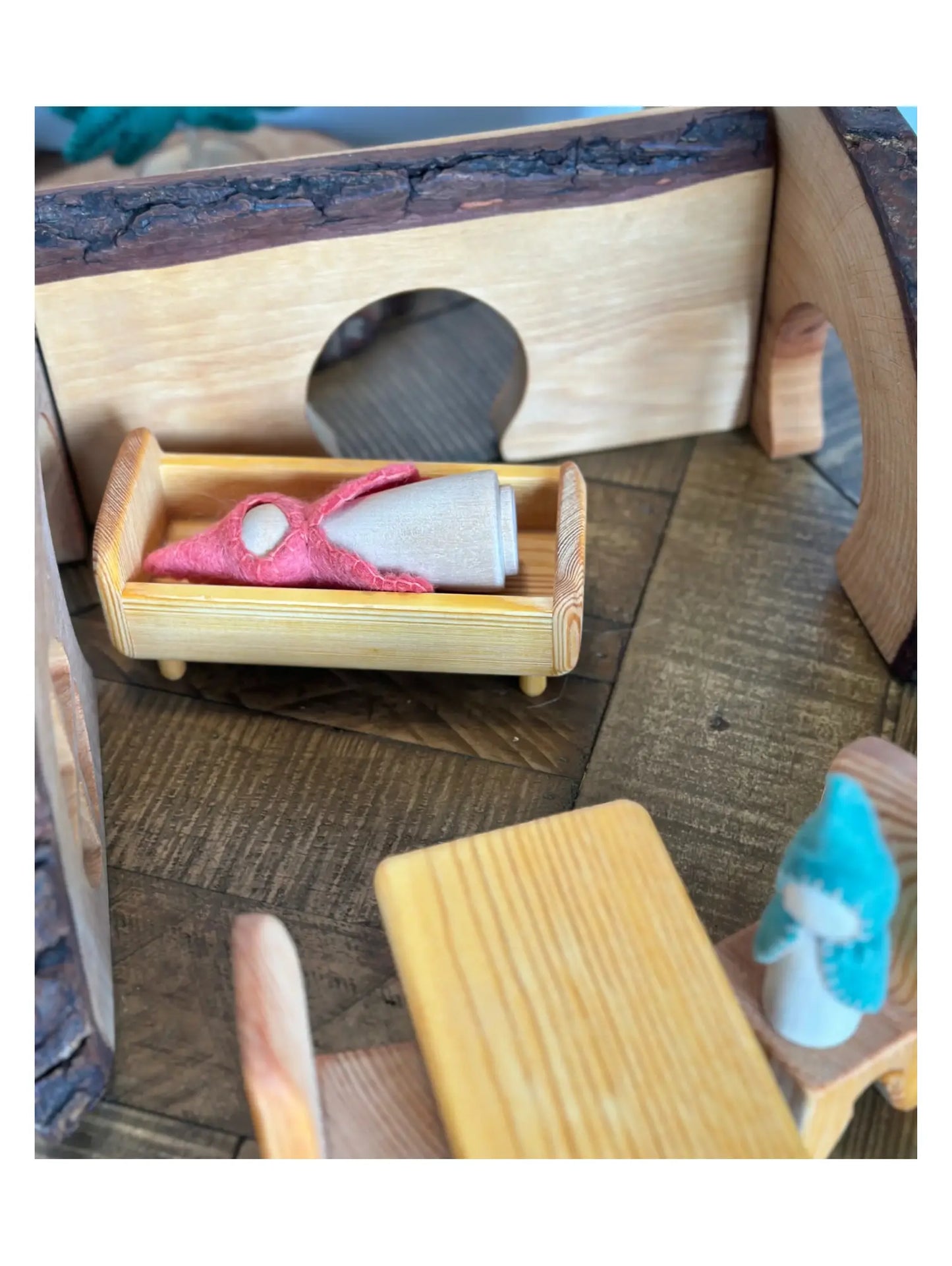 Wooden toy dollhouse bed with a small peg doll figure in a play scene with wooden background