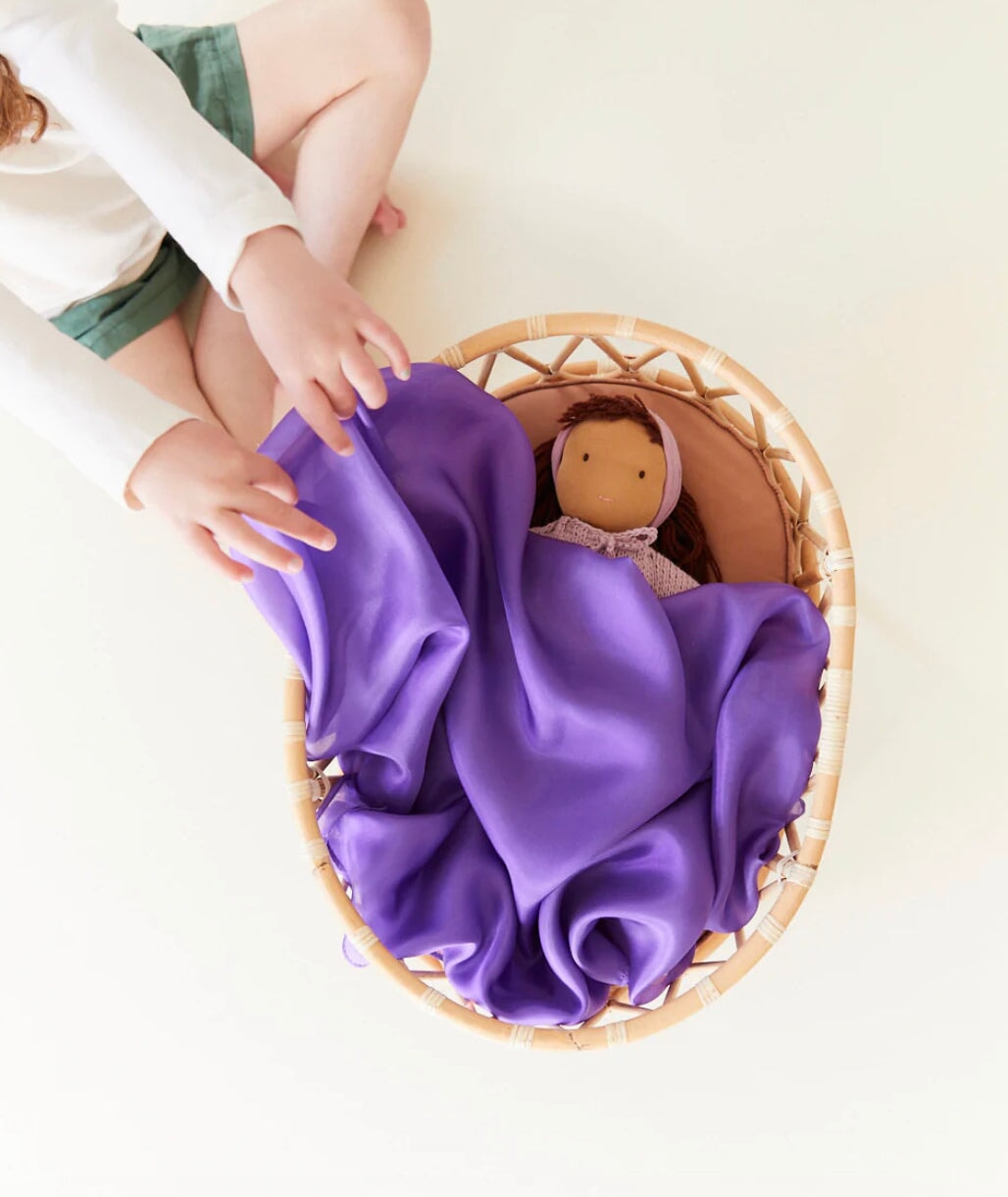 Girl placing a purple playsilk over a doll in a wicker doll bed