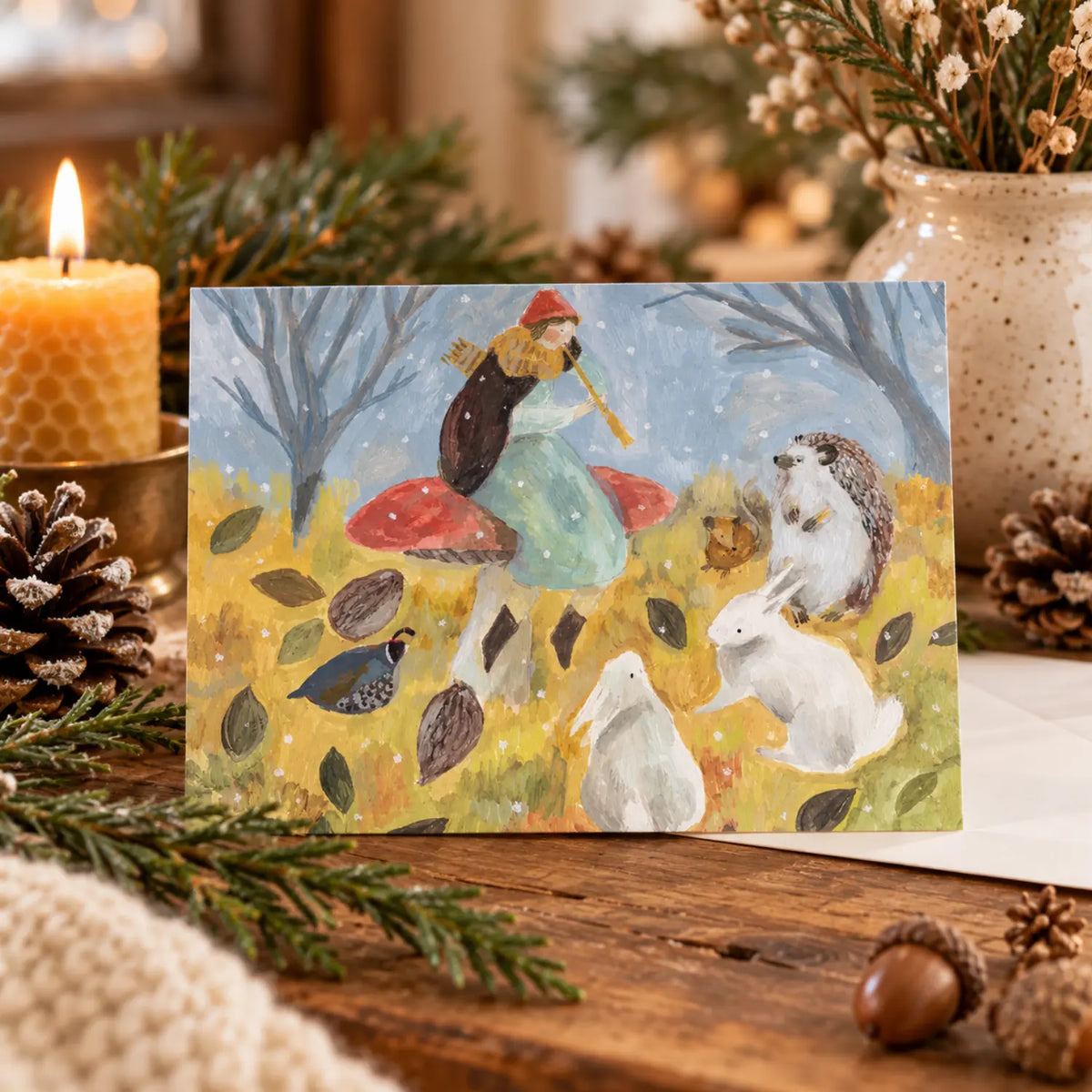 Winter Song woodland postcard styled on a cozy winter nature table with a beeswax candle, evergreens, pinecones and acorns.