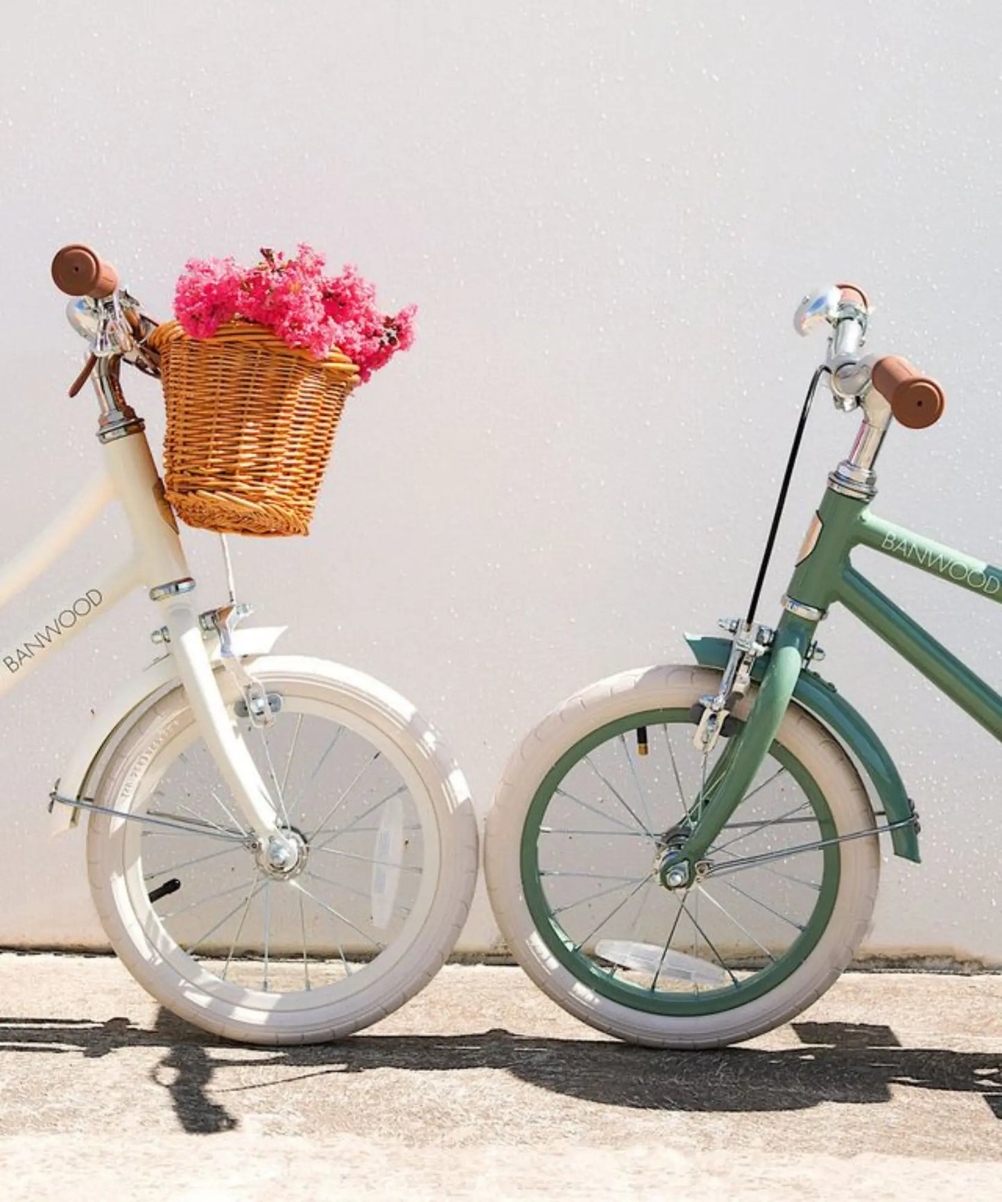 Two children's 20 inch bicycles, one white and one green, with a basket of pink flowers on a light background.