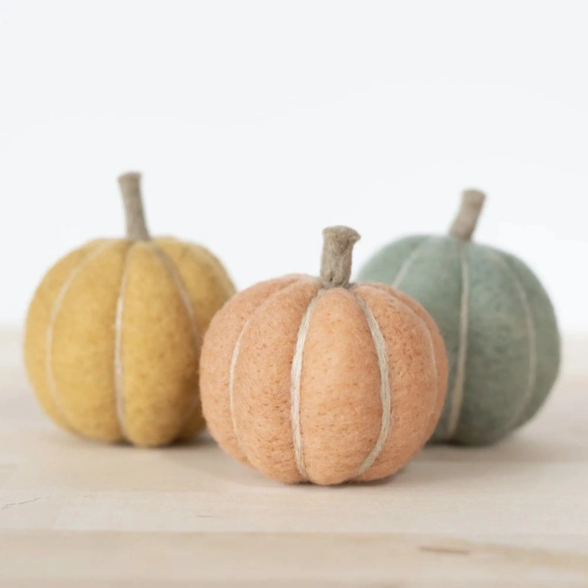 Three needle felted pumpkins in yellow, orange, and green on a wooden surface with a white background