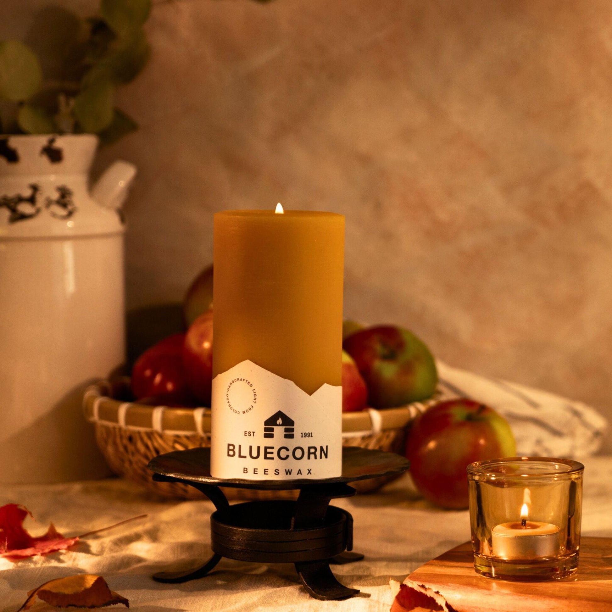 Beeswax candle with Bluecorn label on a rustic setting with apples and a milk churn.