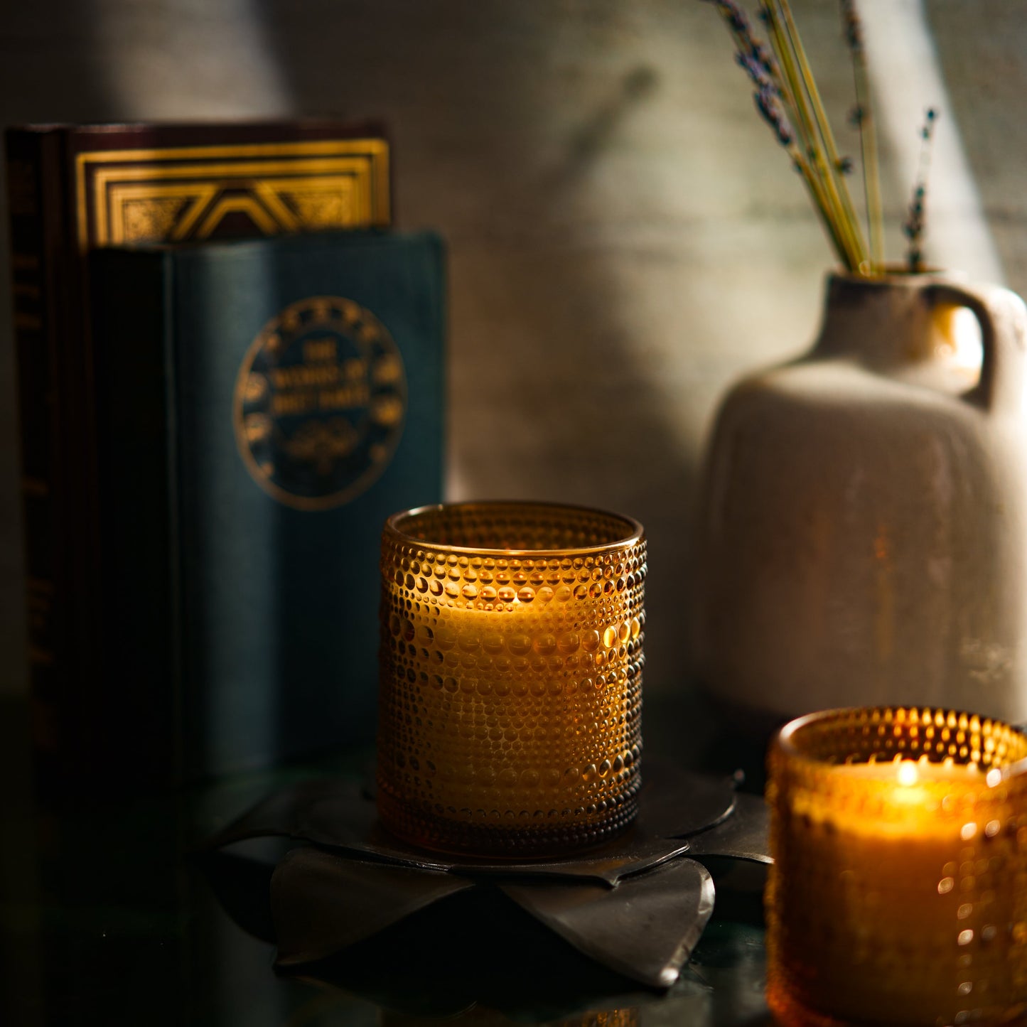 Two lit candles in textured glass holders on a dark surface with a blurred background