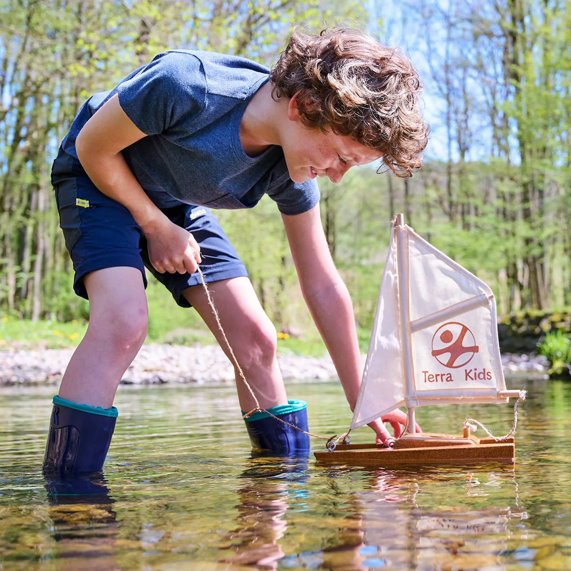 Child playing with a model sailboat in a pond, with 'Terra Kids' branding on the sail.