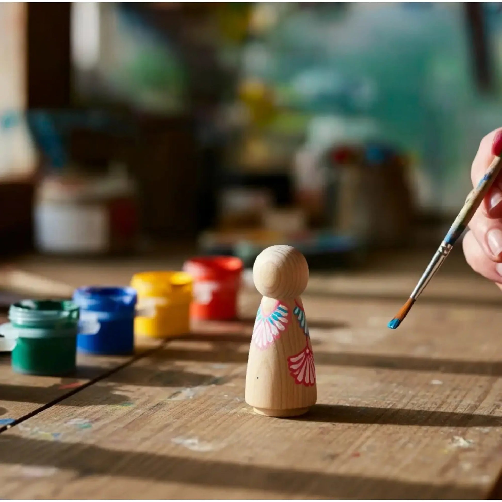 Wooden peg doll with floral design being painted on a wooden table with paint containers.
