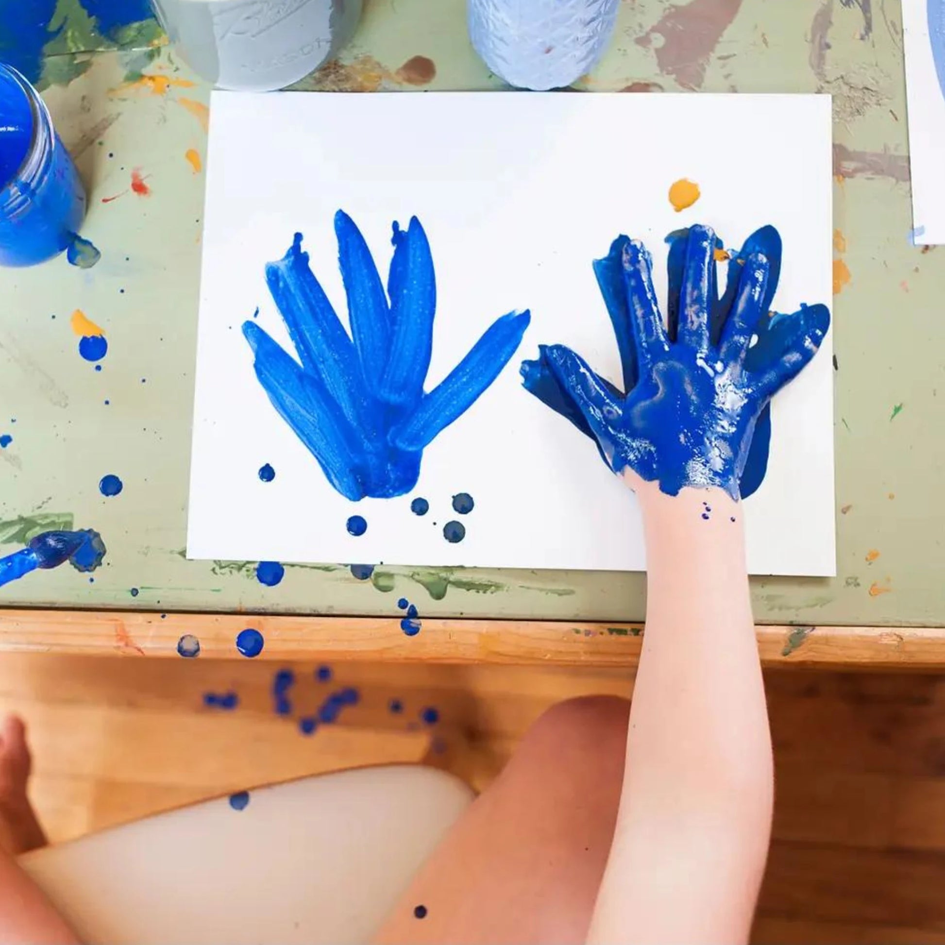 Two blue handprints on a piece of paper with paint splatters around, on a table.