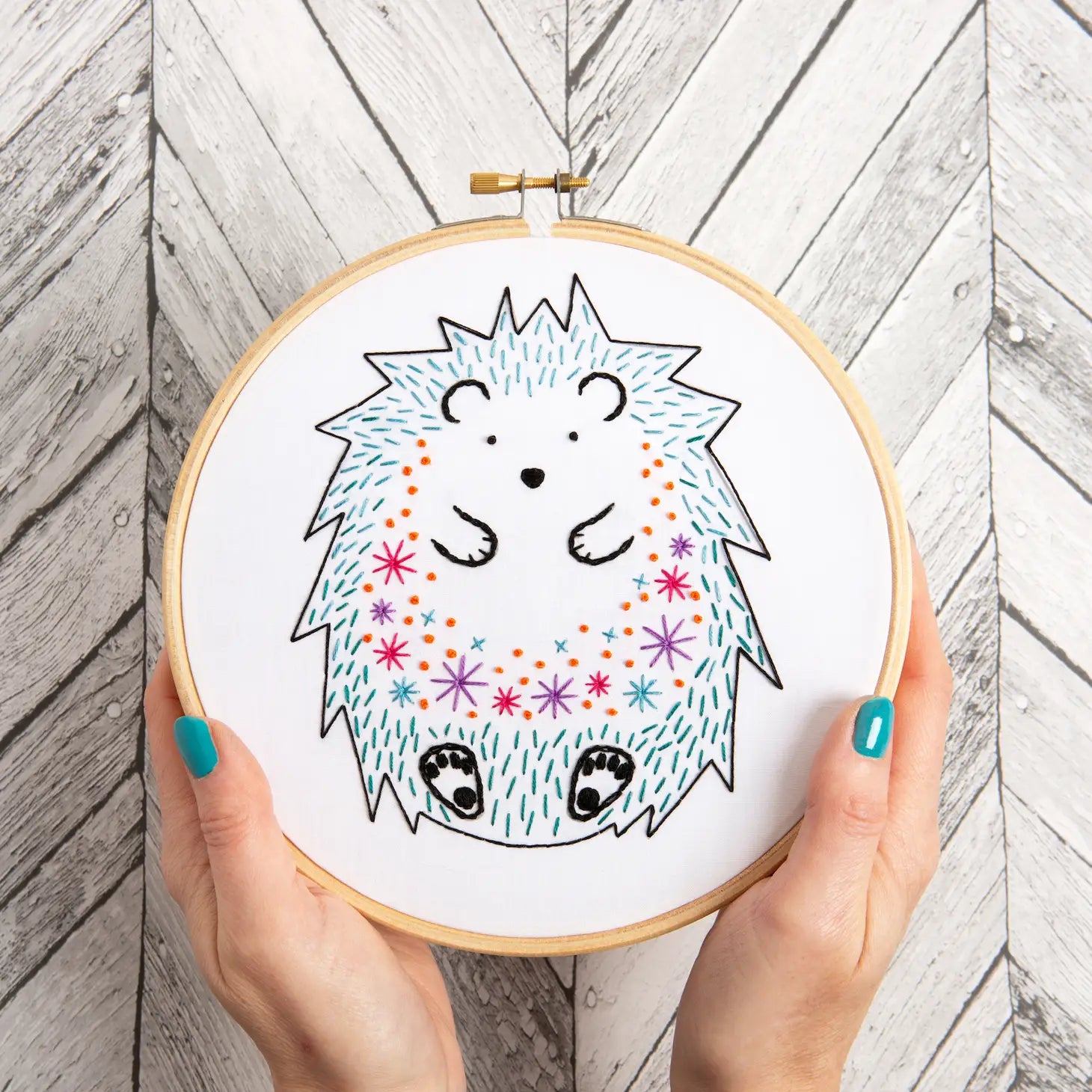 Embroidery hoop with a colorful hedgehog design held by hands on a wooden background