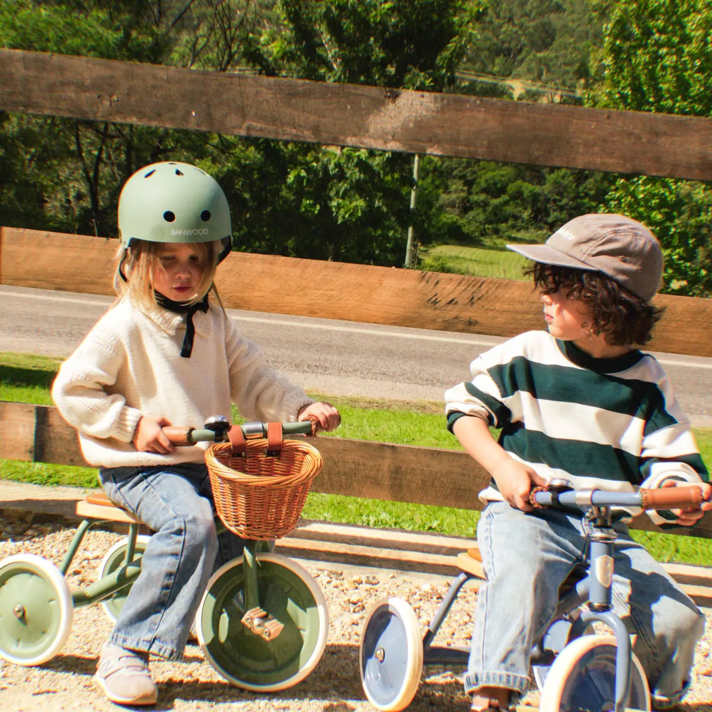 Two children riding vintage Banwood tricycles in forest green and blue in front of a rustic fence