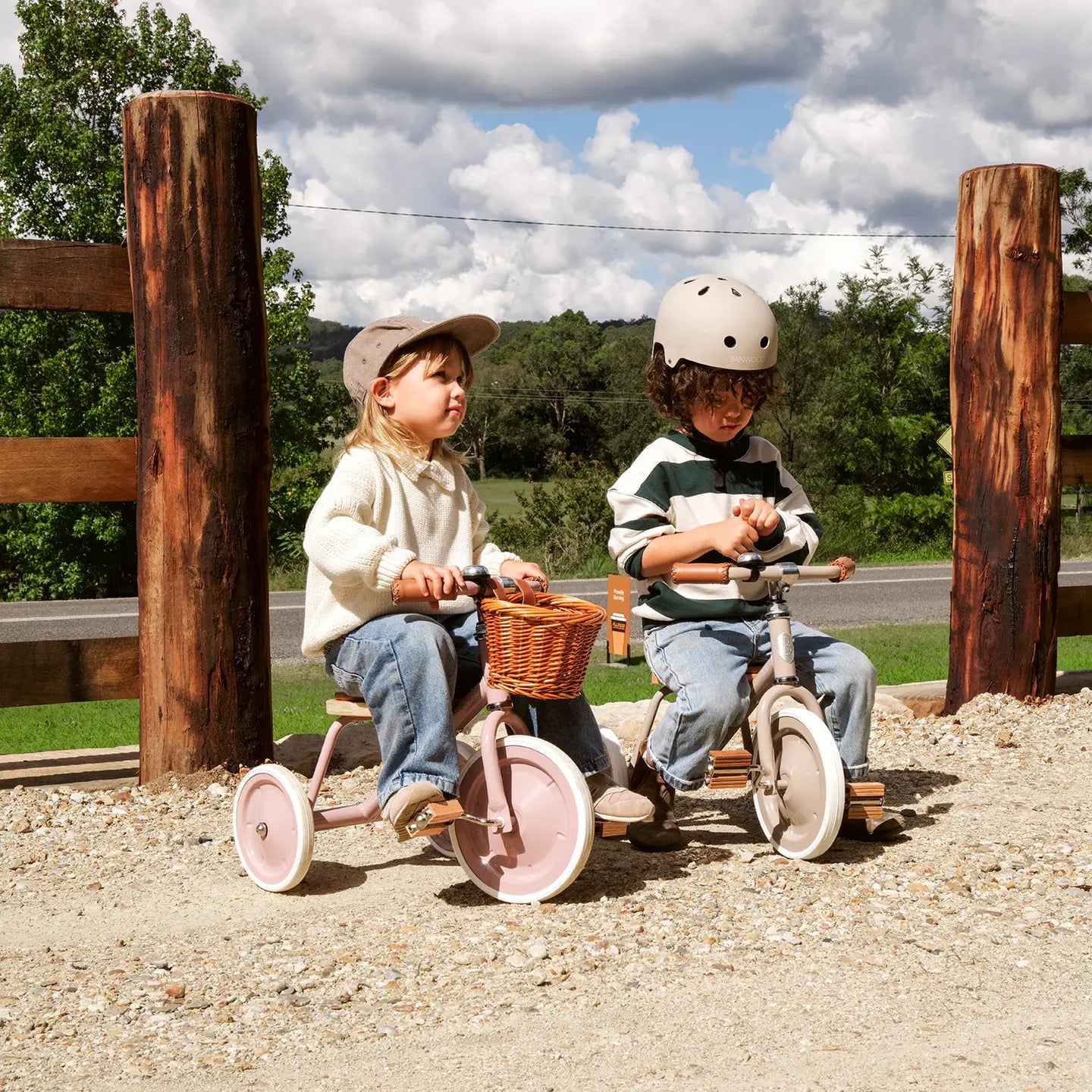 Two children on tricycles in a park setting with wooden posts and greenery.