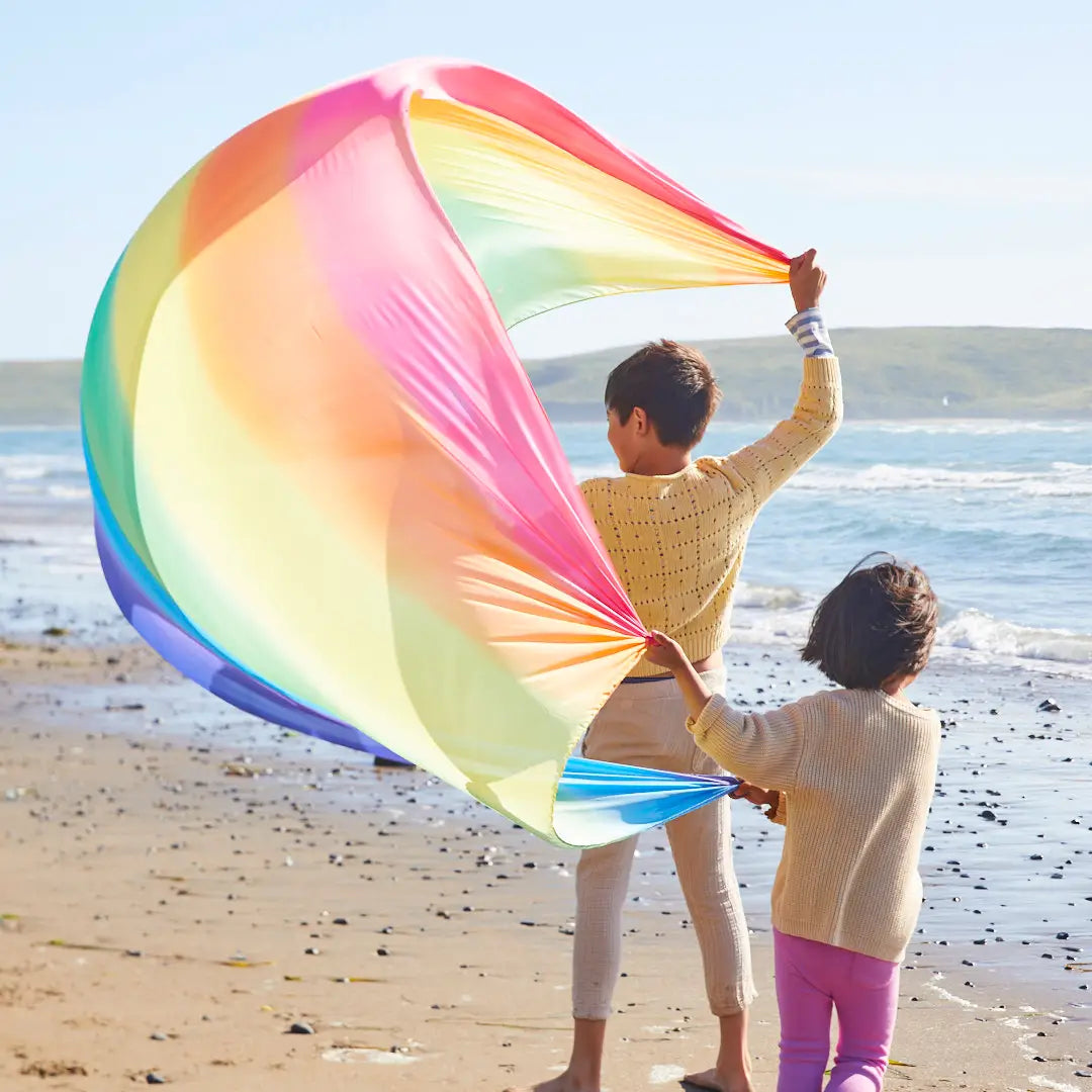 Two children holding a rainbow giant playsilks by Sarah’s Silks on a beach