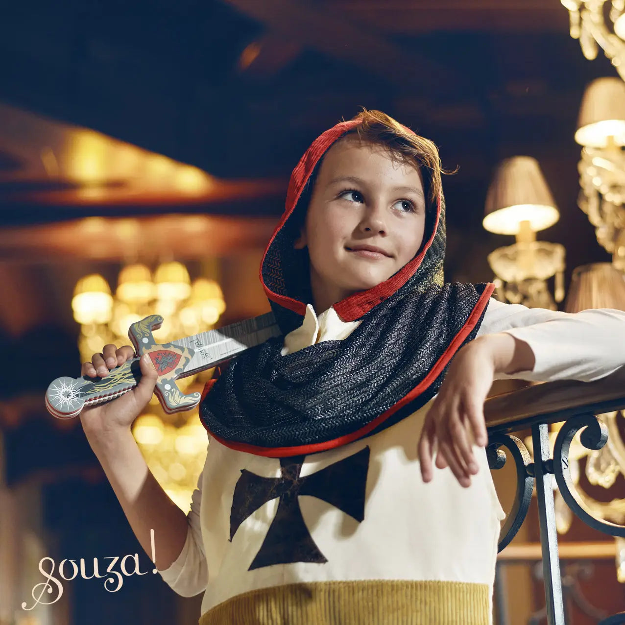Child wearing a Knight's outfit with a chainmail hood and cross design, holding toy swords in an indoor setting.
