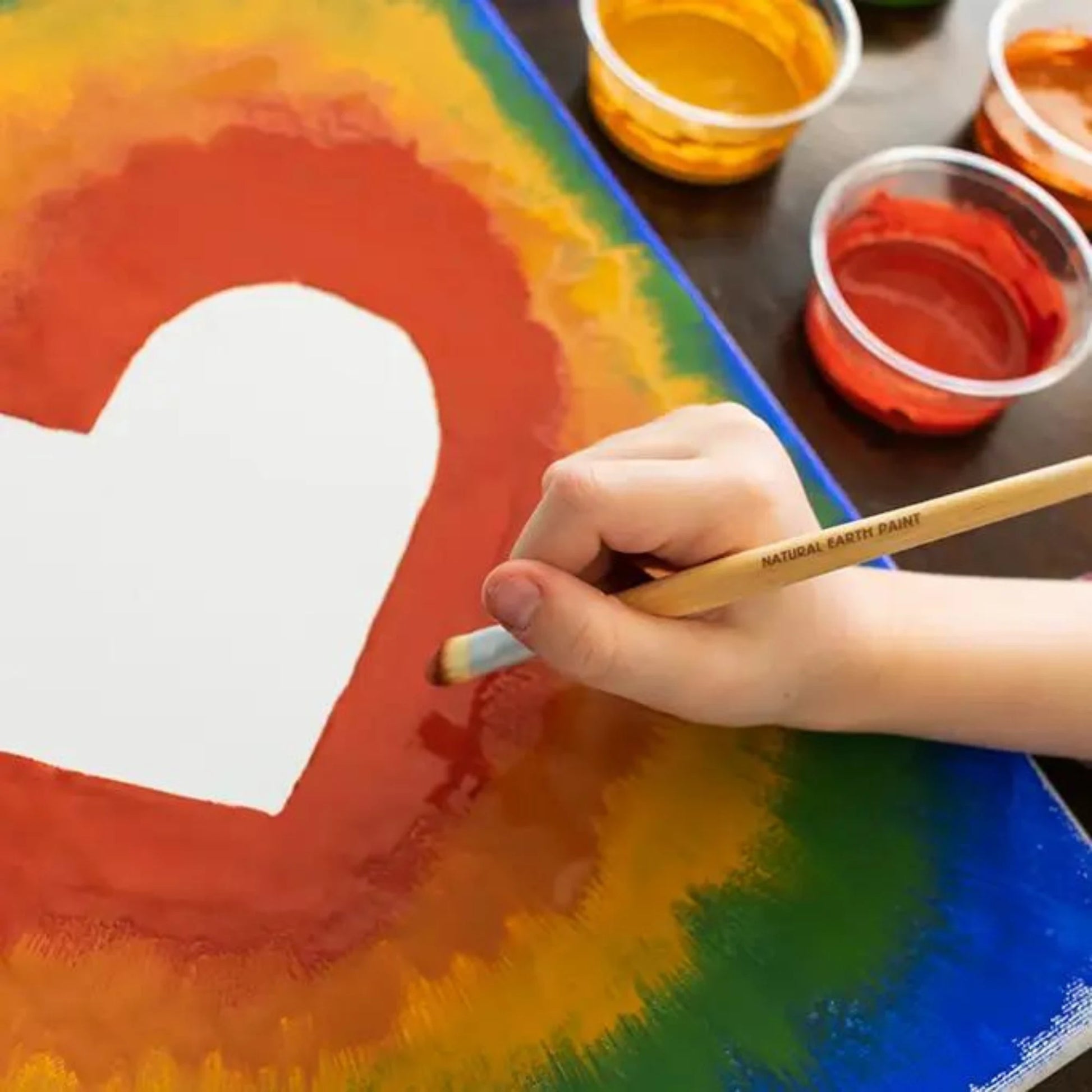 Hand painting a white heart on a colorful watercolor background with paint containers around.
