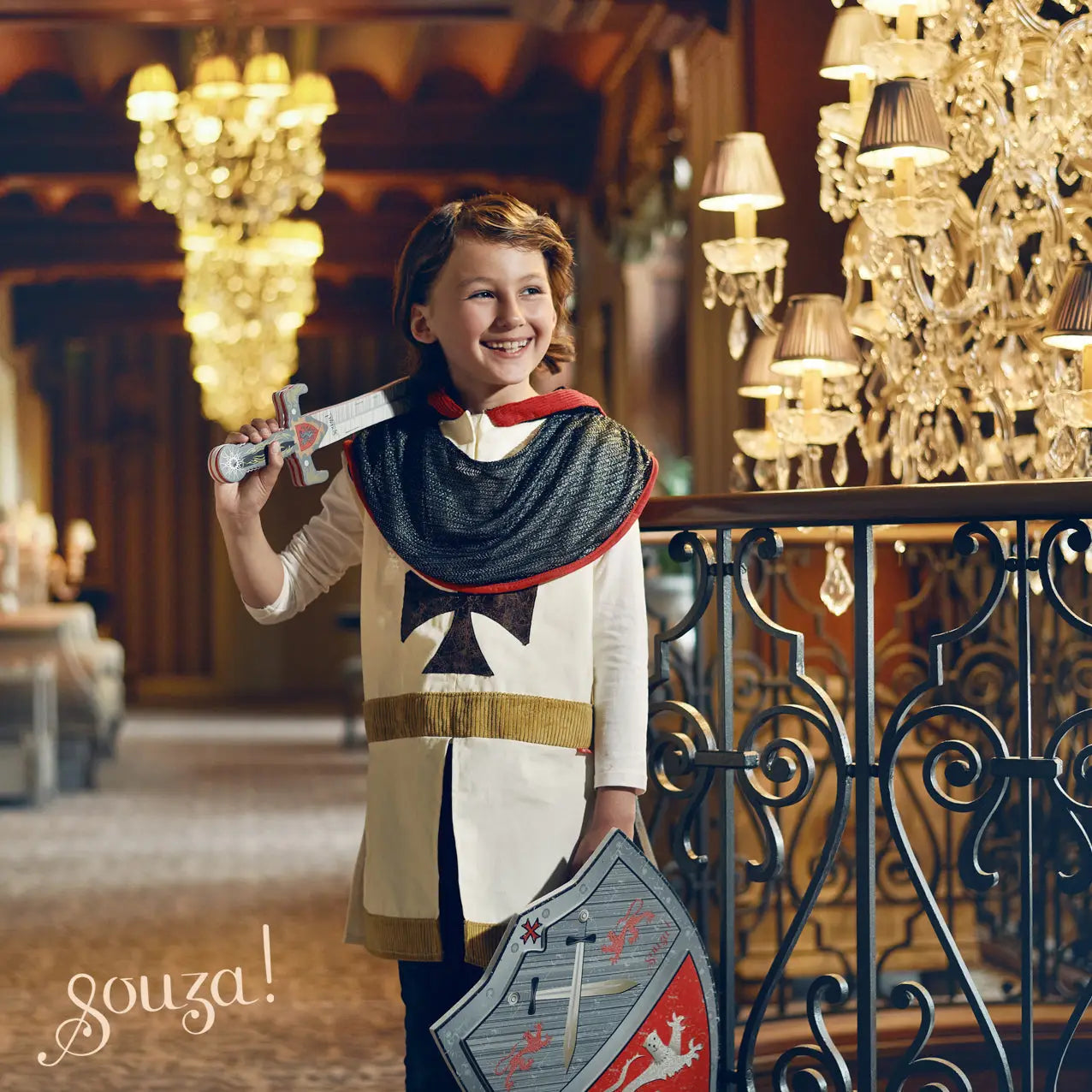 Child in medieval costume holding a sword and shield in an ornate room with chandeliers.