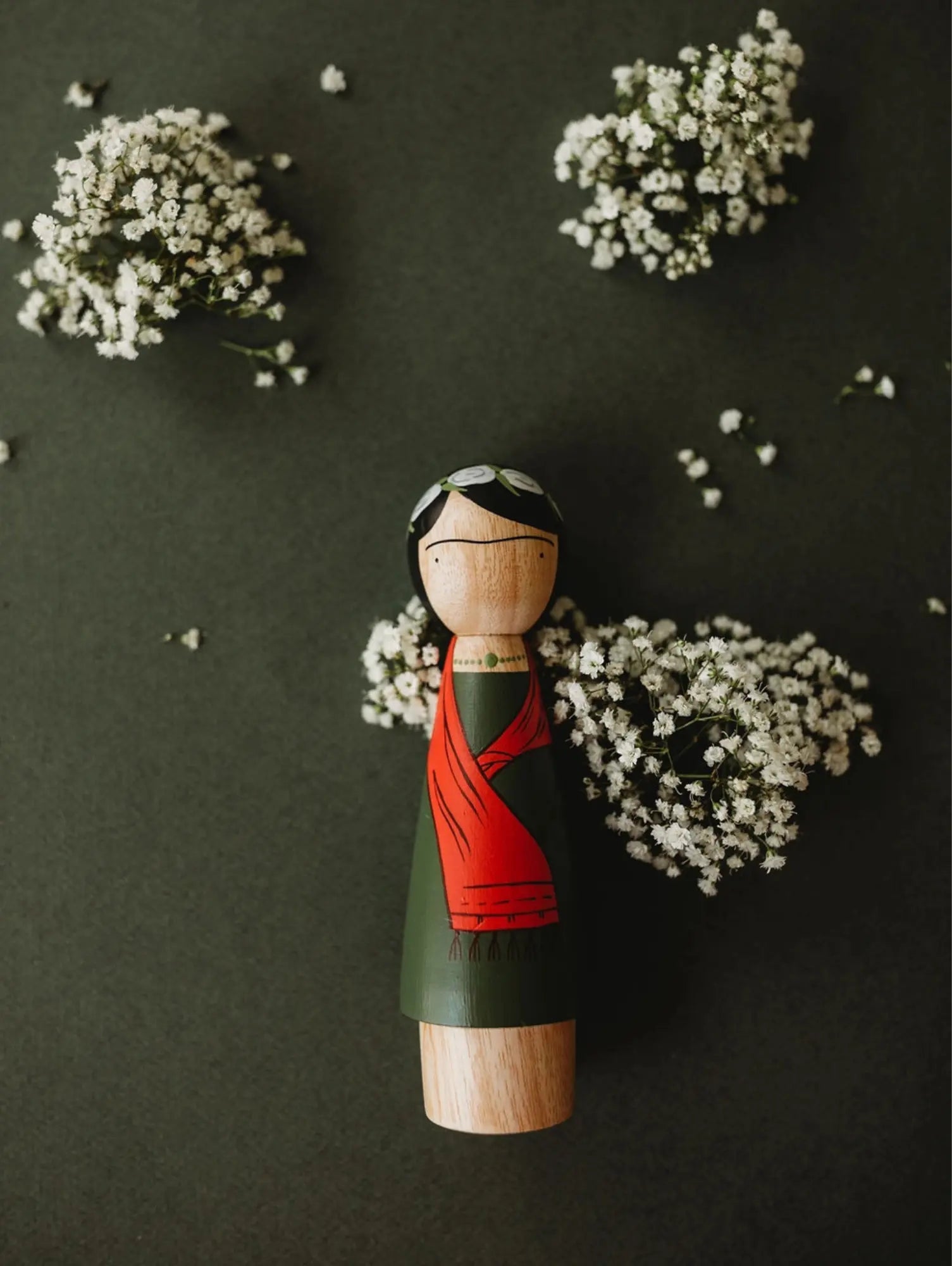 Wooden peg doll with a red and green dress on a dark background with small white flowers.