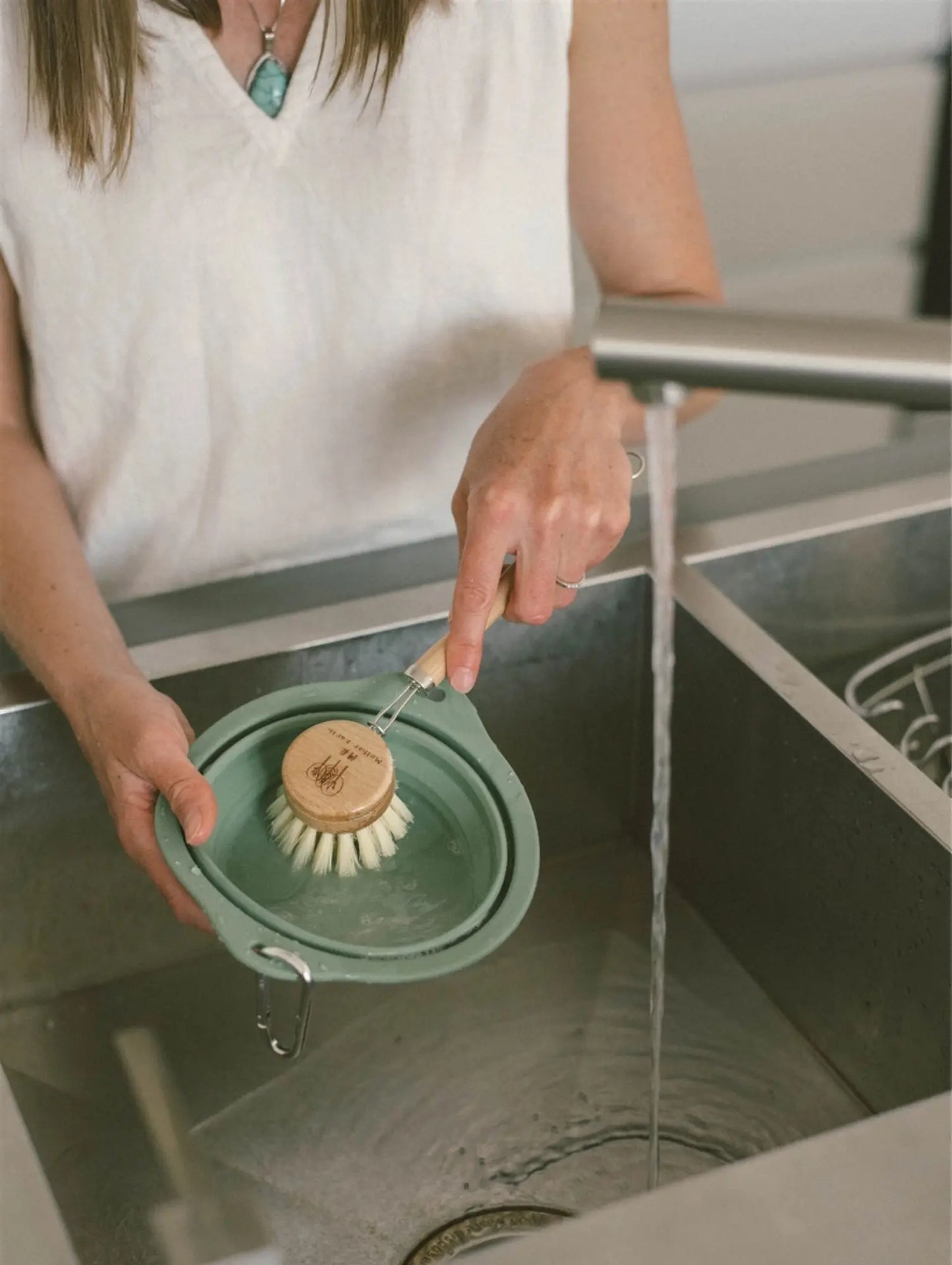 Person washing a green dish brush in a sink with water flowing.
