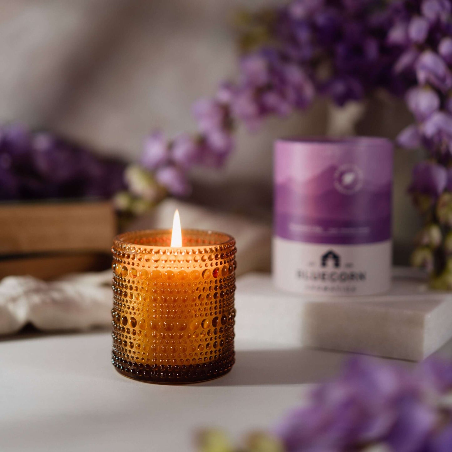 Candle in an amber glass holder with a blurred background featuring purple flowers and a product box.