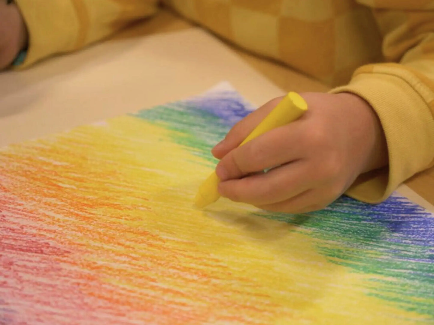 Child's hand holding a yellow crayon over a colorful crayon drawing on paper.
