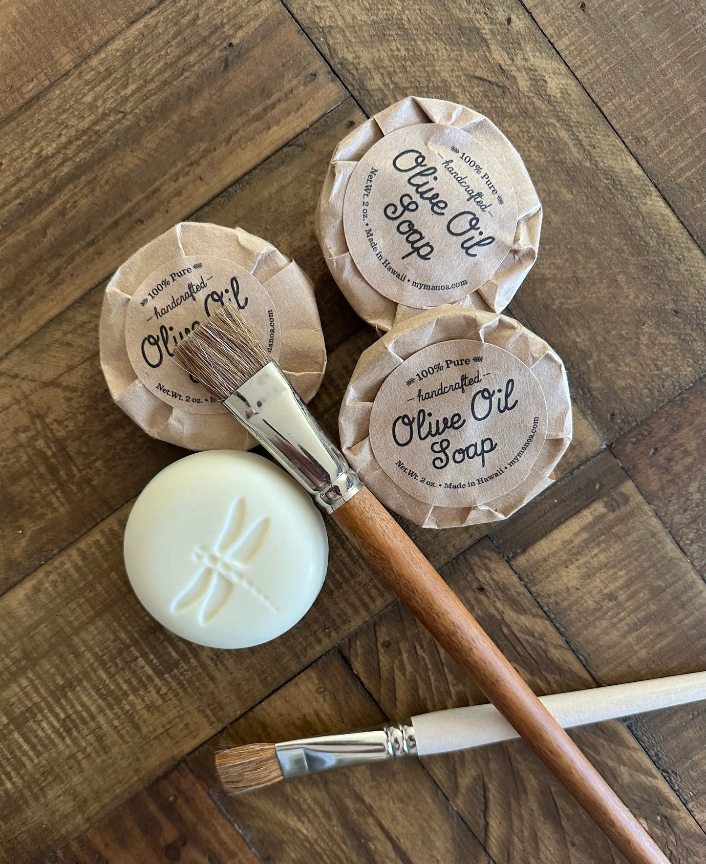 Three bars of olive oil soap with labels on a wooden surface, accompanied by two paintbrushes.