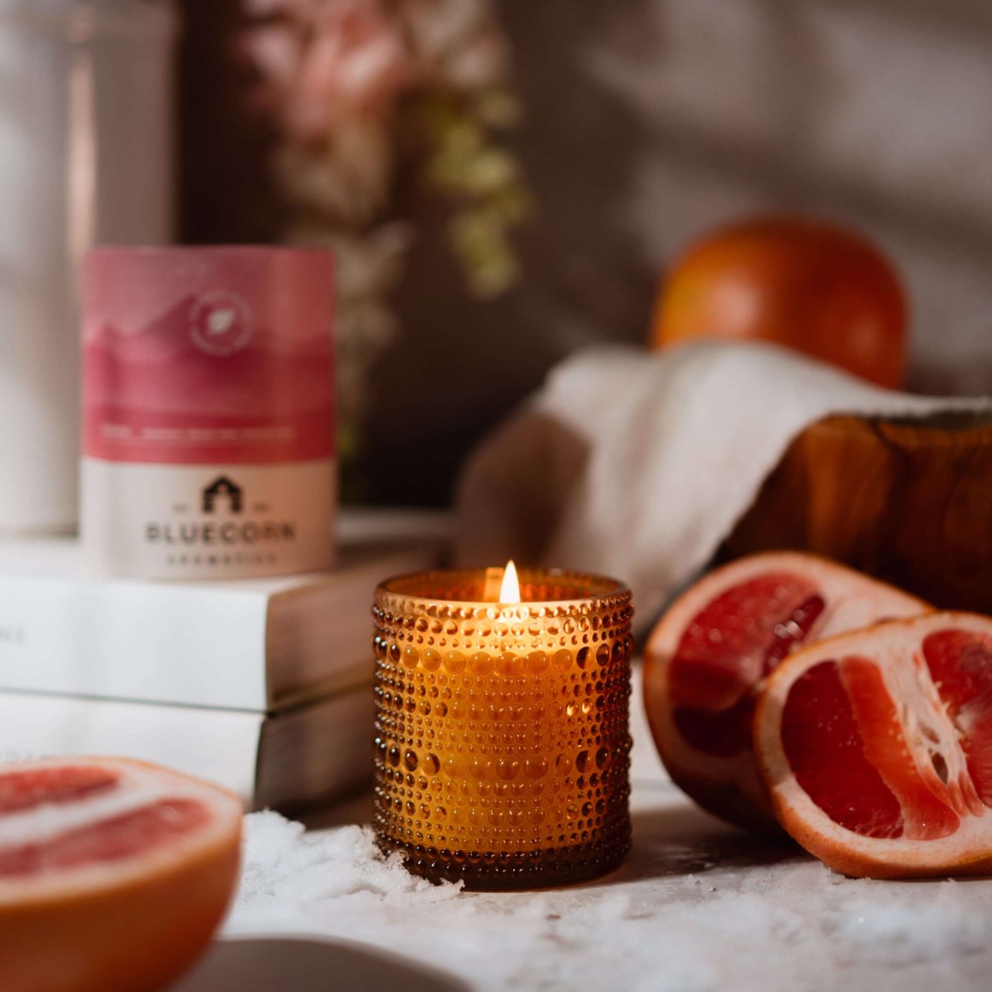 Candle in a textured holder with grapefruits and a product labeled 'Bluecorn' in the background.
