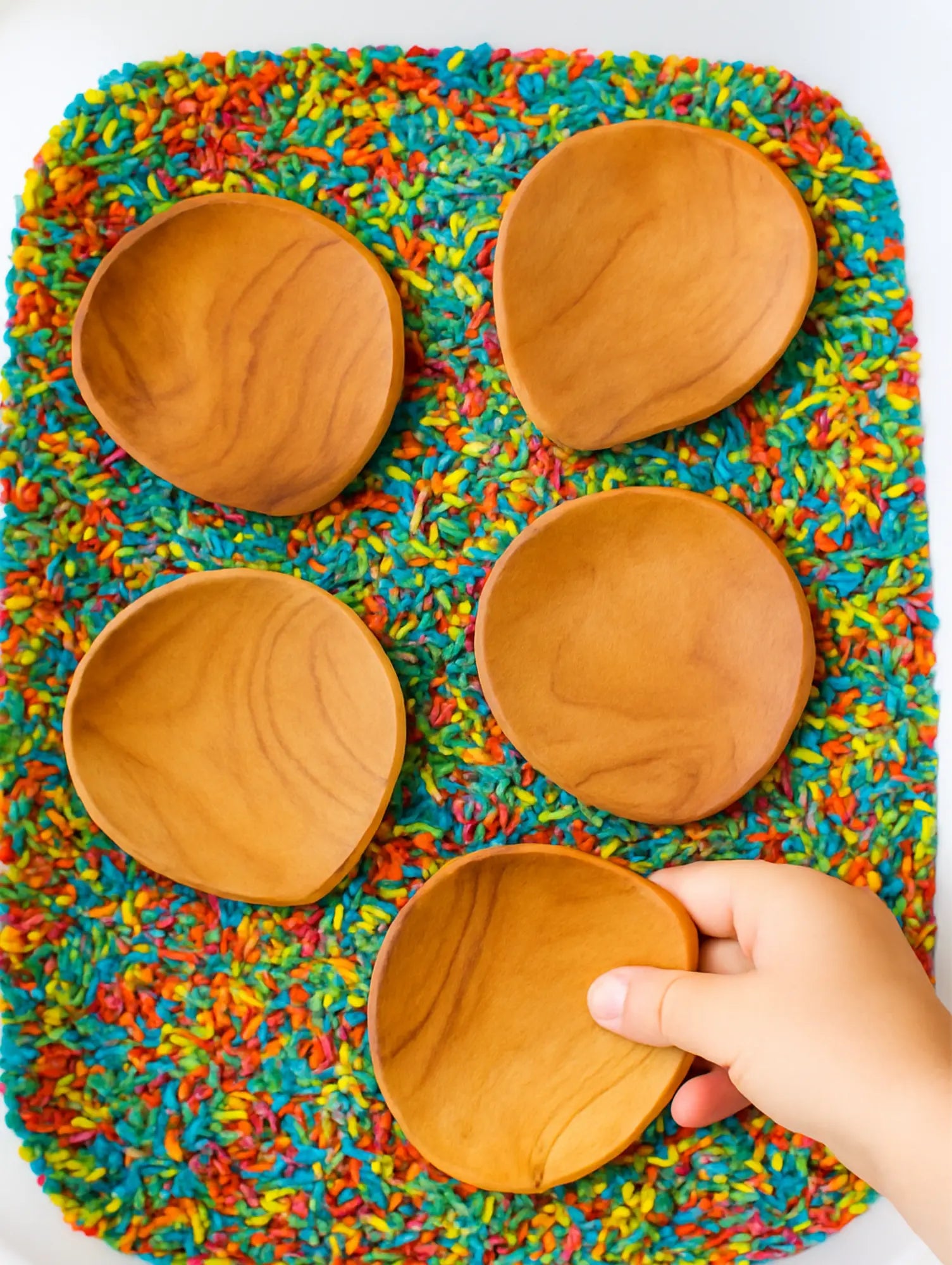 Four wooden scrapers in a colorful rice filled sensory bin with a hand holding one 