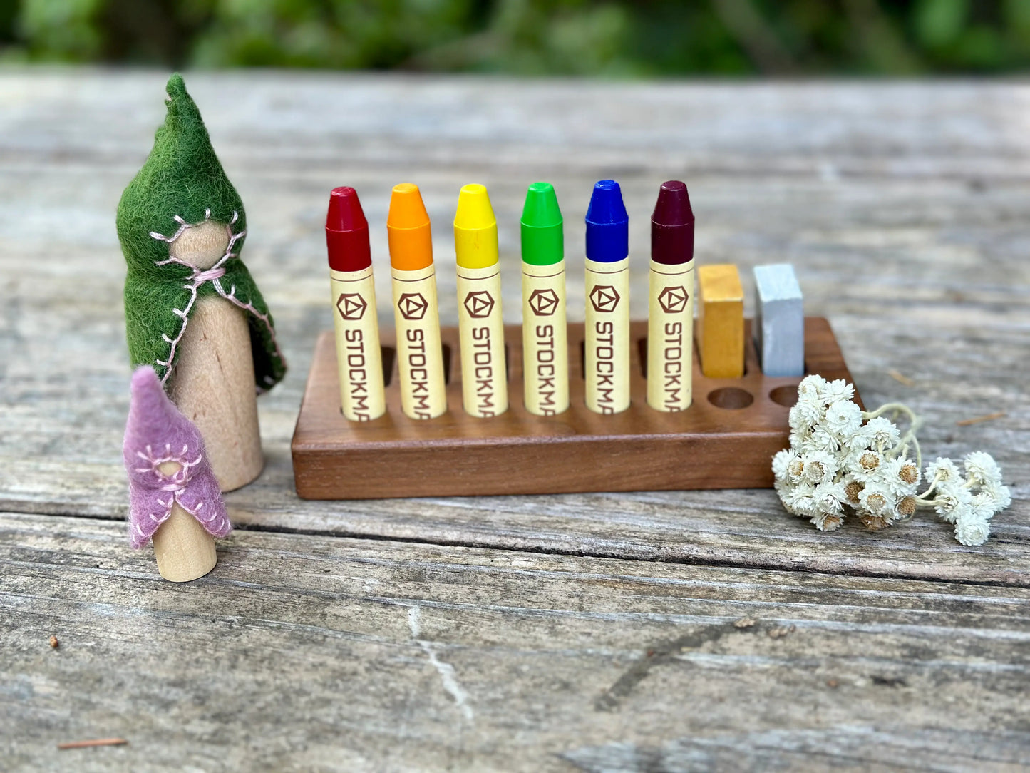 Colorful Stockmar crayons with wooden holder on a wooden table, with small figures and flowers.