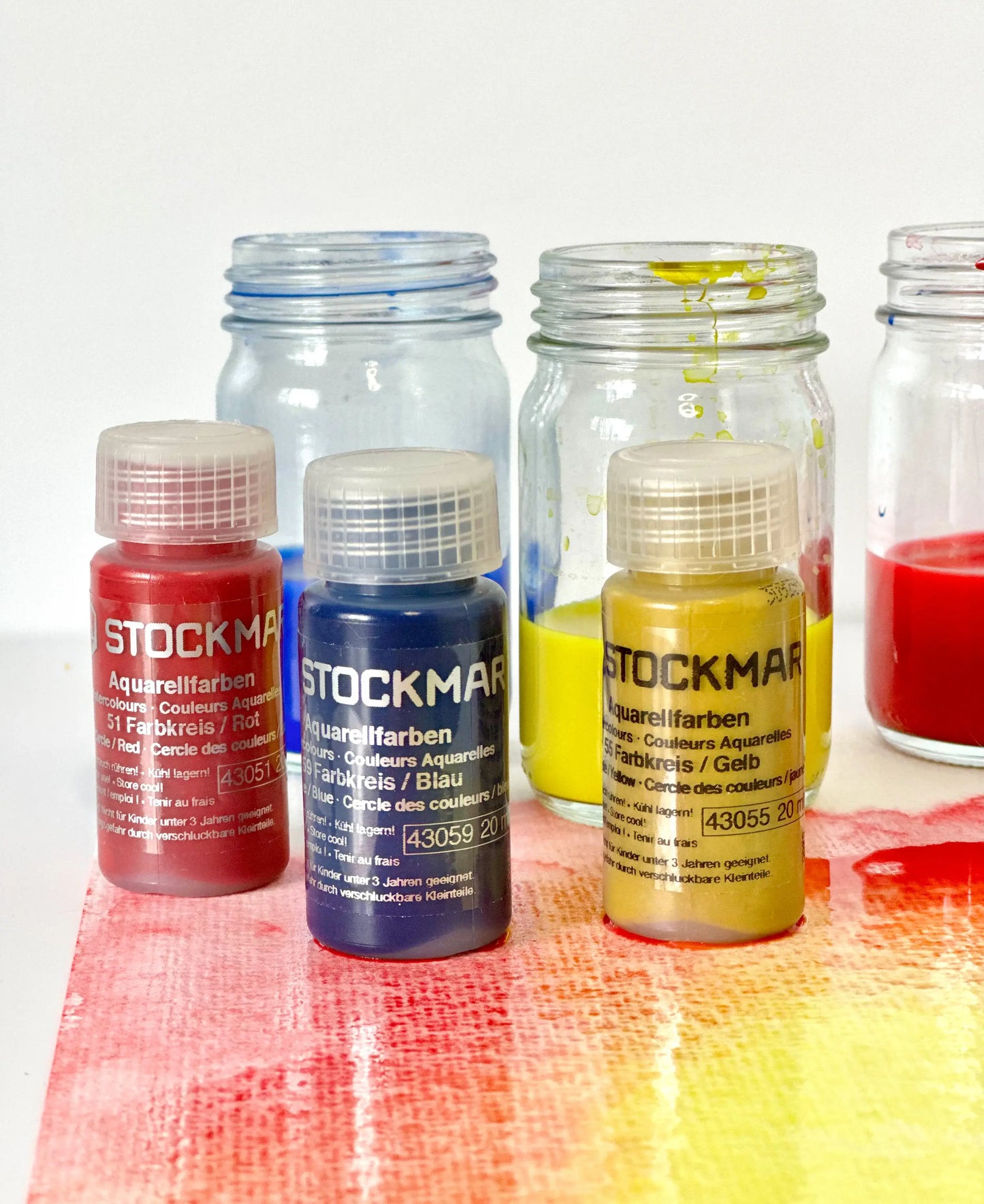 Set of Stockmar watercolor bottles on a colorful surface with jars in the background.