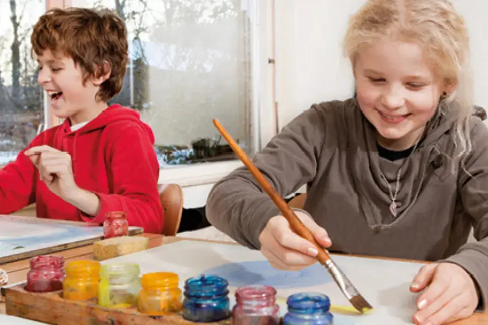 Two children sitting at a table with paint jars and brushes, engaged in an art activity.
