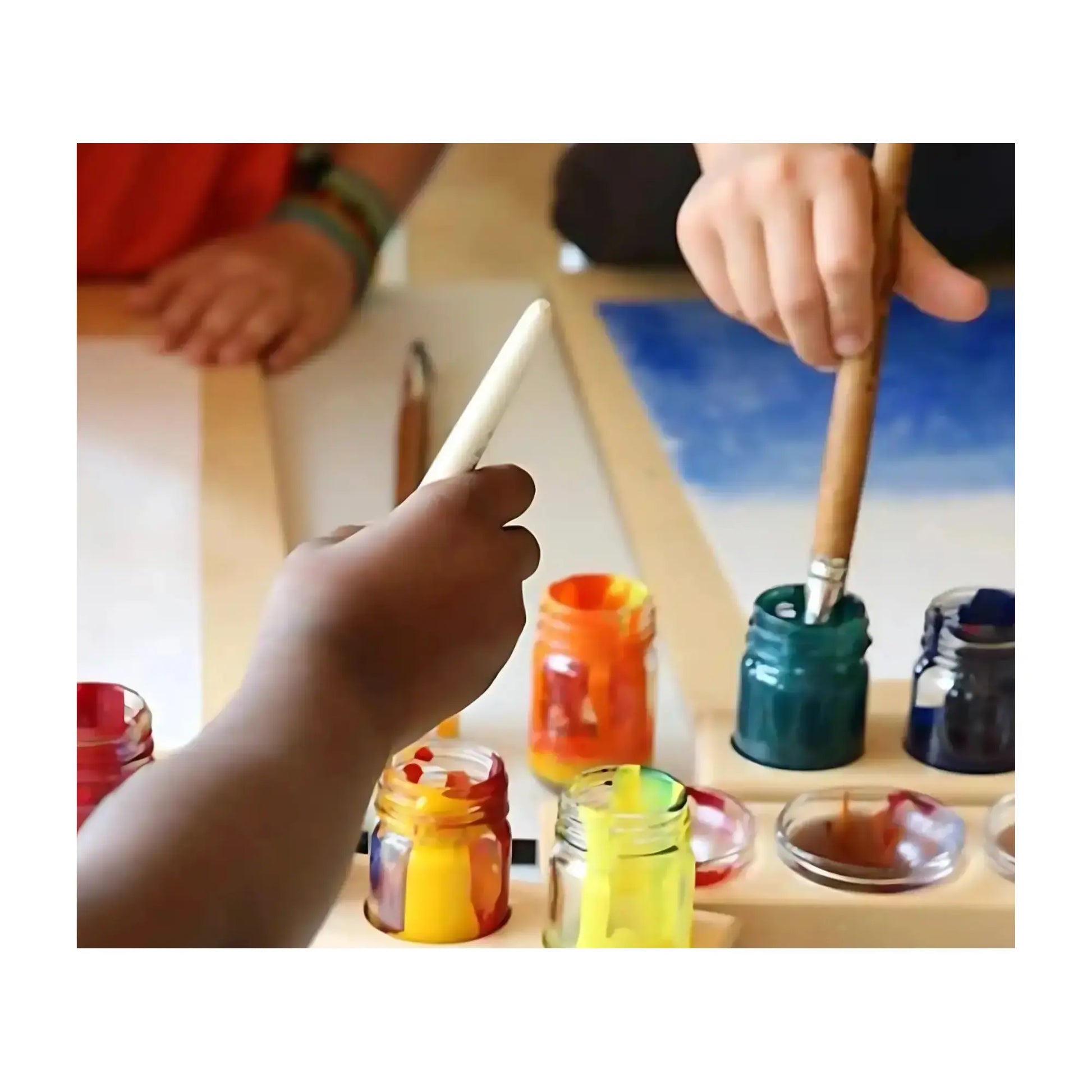 Children's hands with paintbrushes and colorful paint containers on a table.