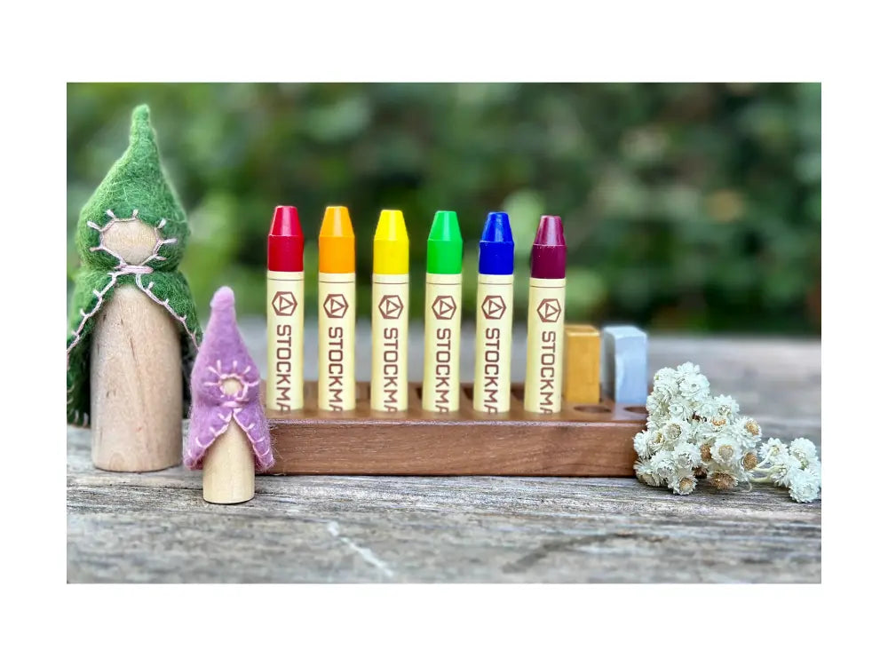Set of colorful Stockmar crayons on a wooden stand with small figurines and flowers in the background.