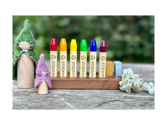 Set of colorful Stockmar crayons on a wooden stand with small figurines and flowers in the background.