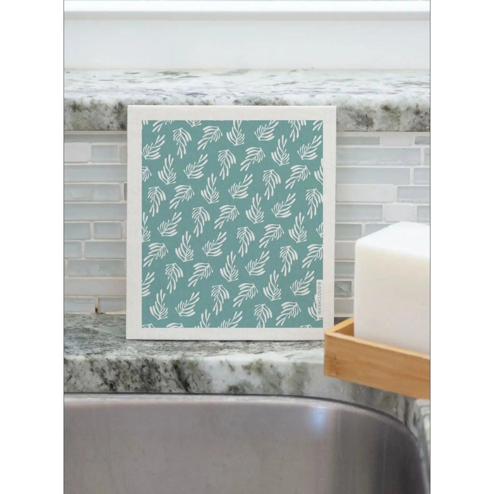 Swedish Dishcloth in Teal and White with Kelp Design Propped up against the kitchen sink backsplash