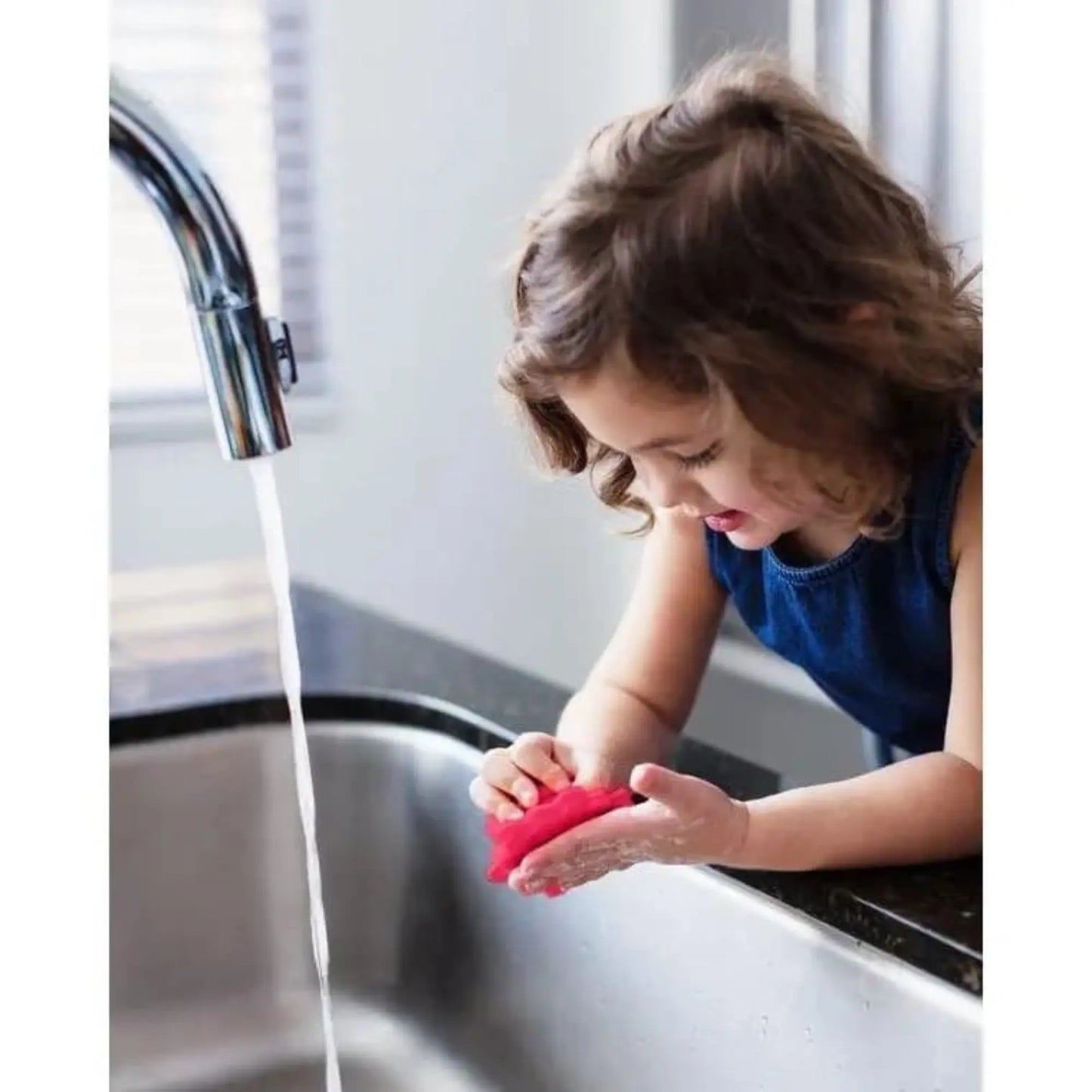 Child washing hands with a red silicone scrubby in a kitchen sink.