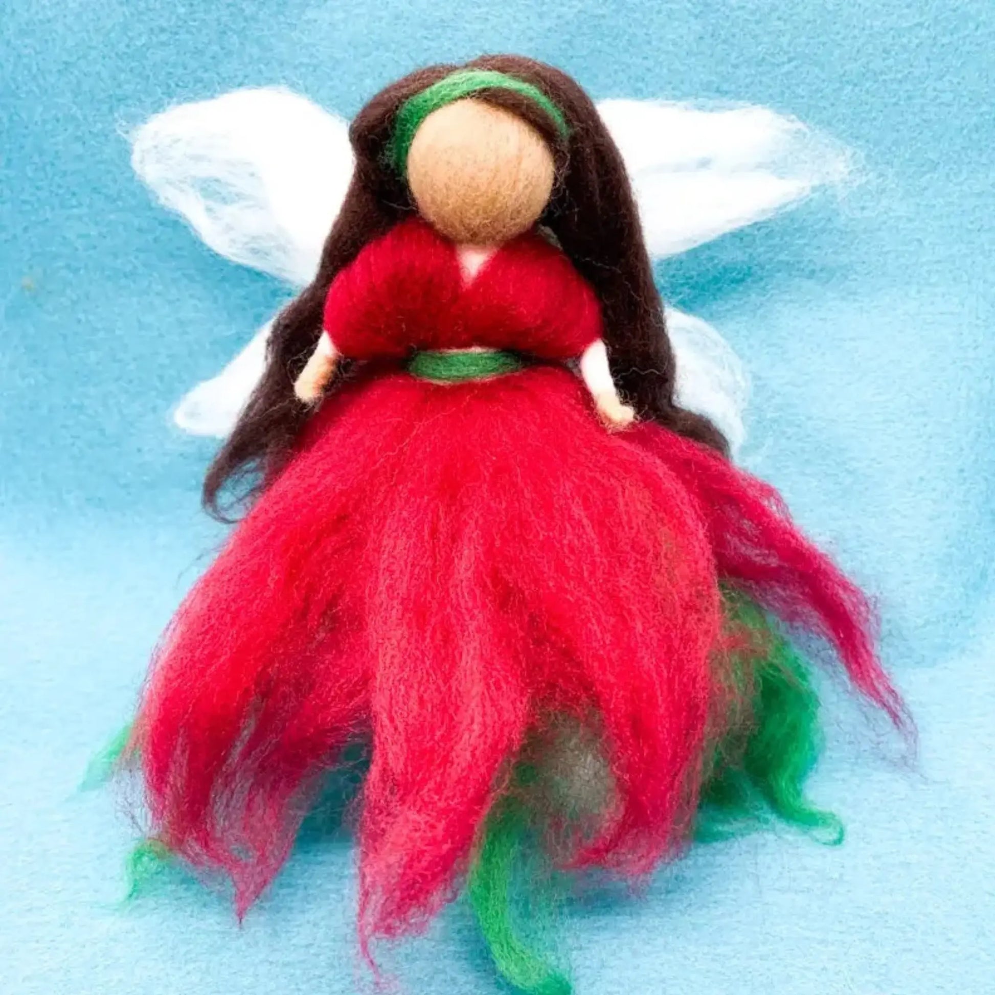 Felted fairy doll with white wings, red and green dress, and brown hair on a blue background