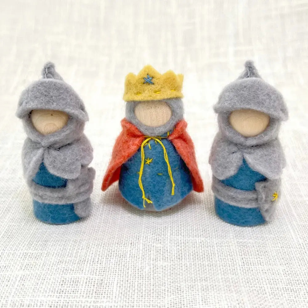 Three felted dolls dressed in blue and gray with a red cape and yellow crown on a textured white background.