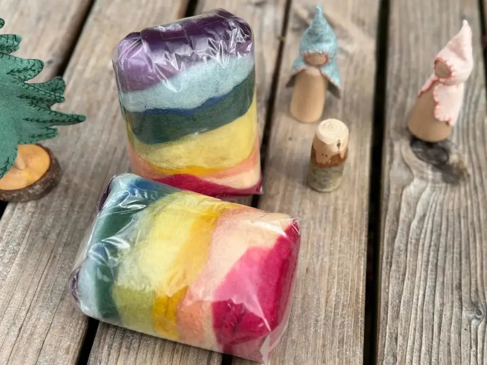 Two bags of colorful plant dyed wool felt on a wooden table with peg dolls and a felt tree