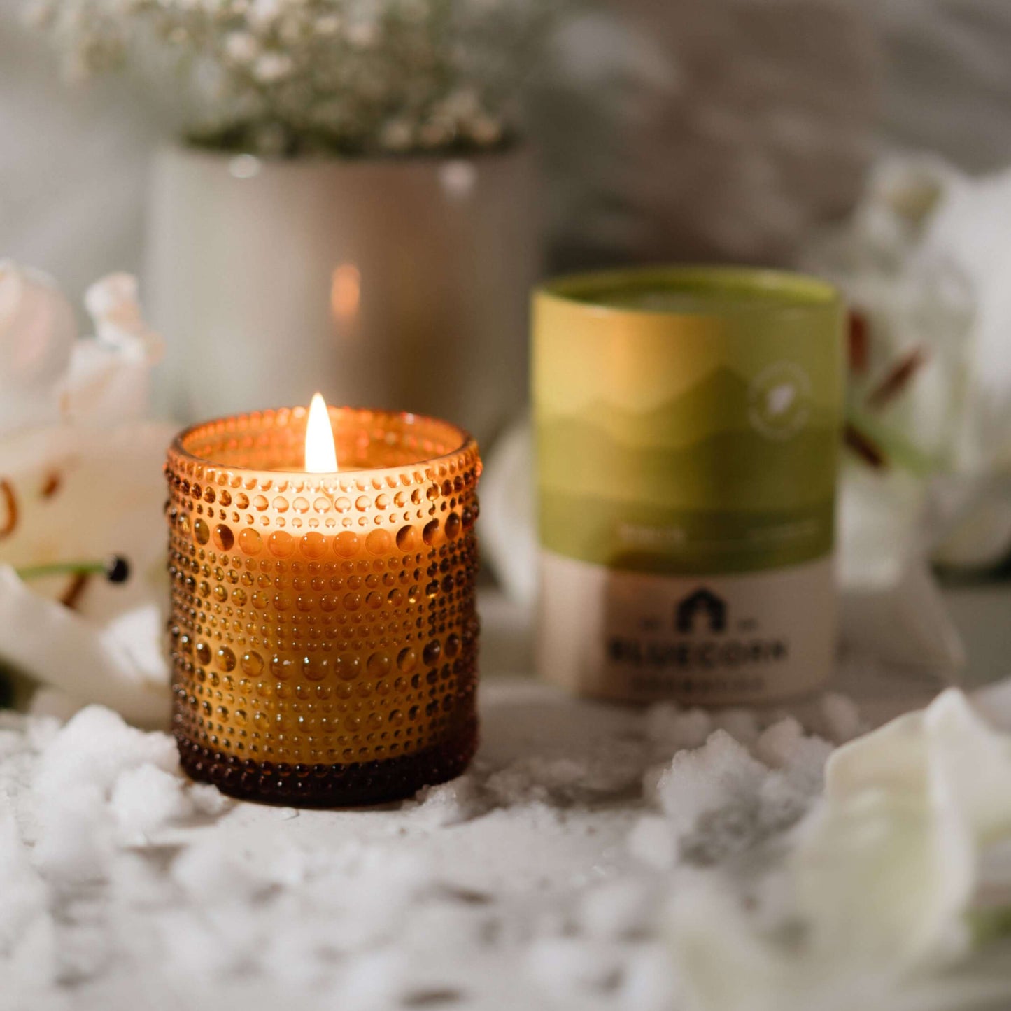 Candle in a textured holder with a blurred background of flowers and a product box.
