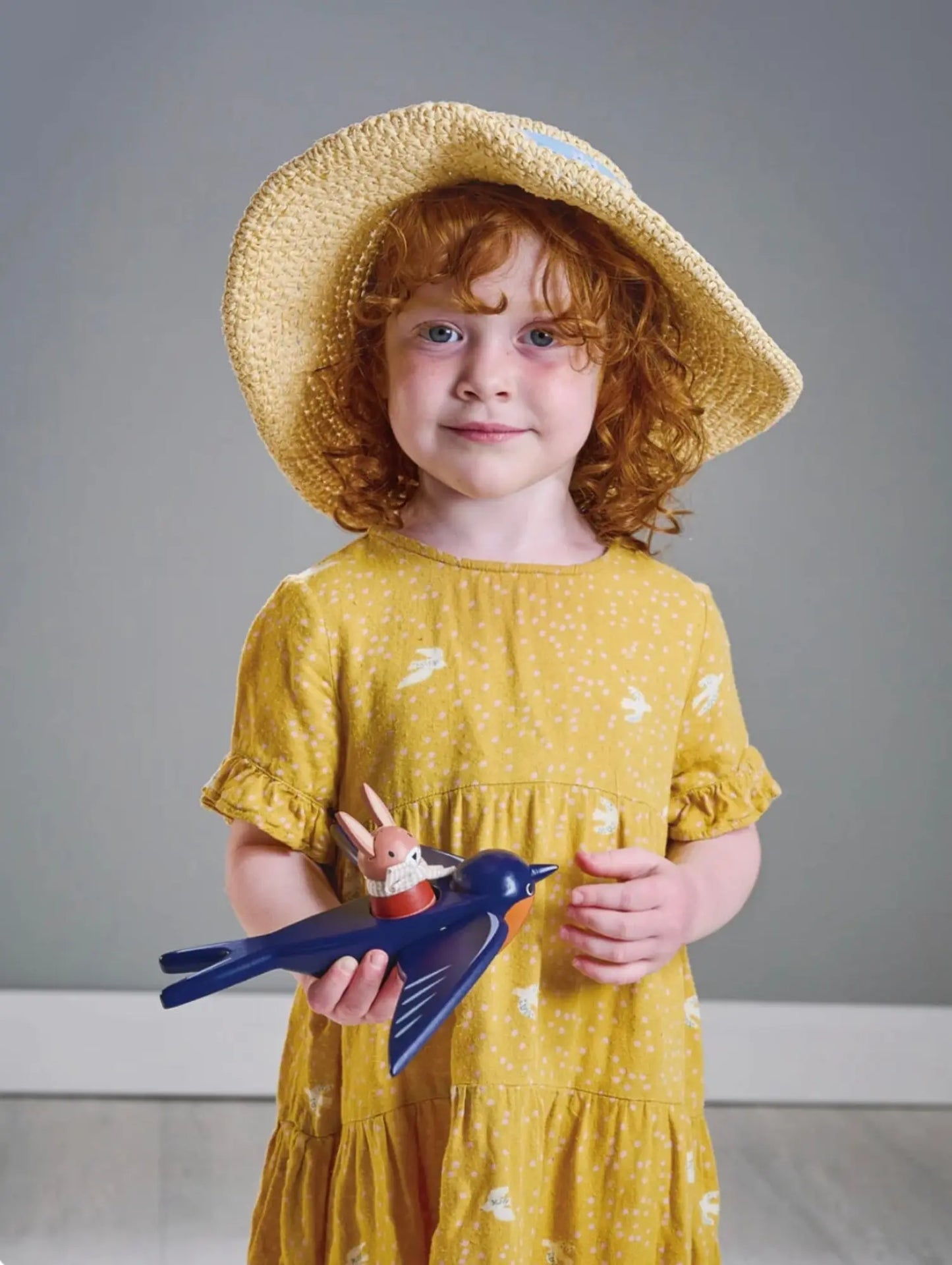 Young girl in a yellow dress and straw hat holding a toy bird and hare against a gray background