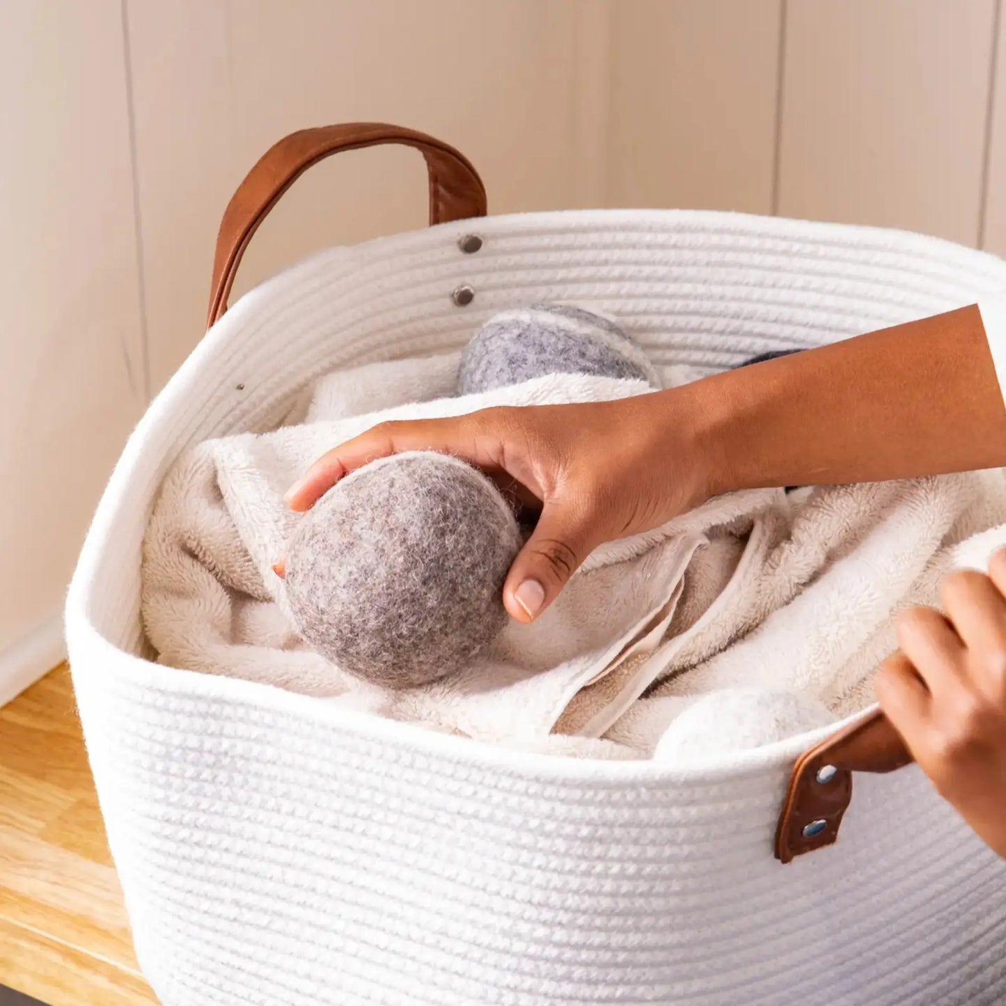 Person placing a gray dryer ball into a white laundry basket with brown handles.